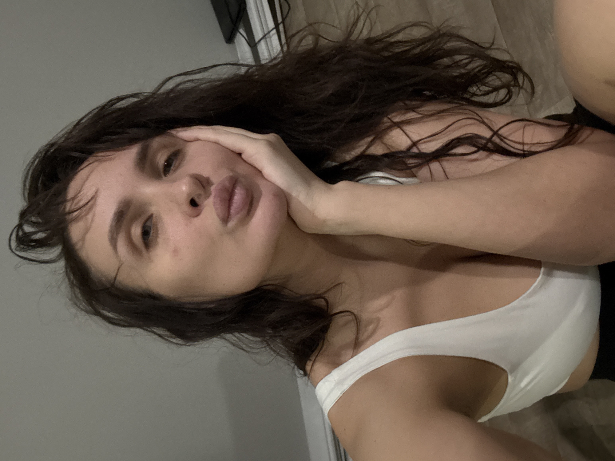 A woman with brown hair and fair skin is lying on her side. She is wearing a white tank top. Her left hand is supporting her face as she pouts her lips towards the camera. The background is a plain wall. The image location is not discernable from the available information.