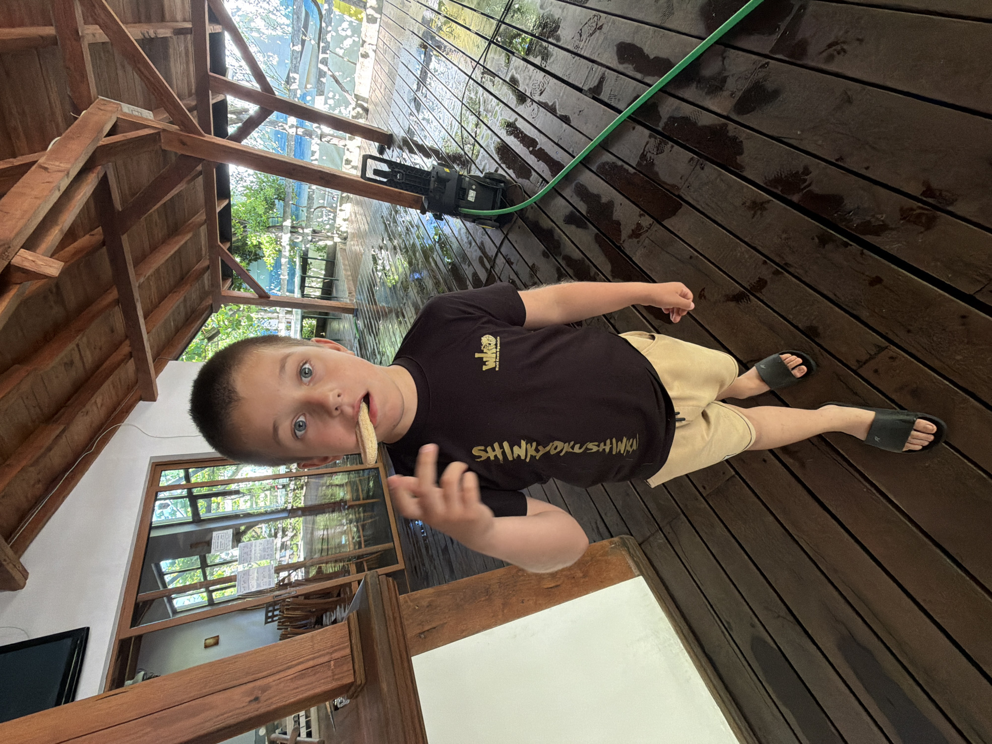 The image shows a young boy with blue eyes and short brown hair, wearing a black t-shirt with gold lettering that reads "SHINKYOKUSHINKAI" and a small logo, along with tan shorts and black slides. He is standing on a wet wooden deck, holding a piece of food in his mouth and with his right hand raised. To the right of the boy, a green garden hose is connected to a black pressure washer. The deck appears to be part of an outdoor covered area with a wooden roof structure. In the background, there are glass doors and windows reflecting the outdoors, and glimpses of greenery and water. The scene is located in Tsarevo, Bulgaria, likely at a restaurant or resort with an outdoor seating area. The wet deck suggests recent cleaning or rain.