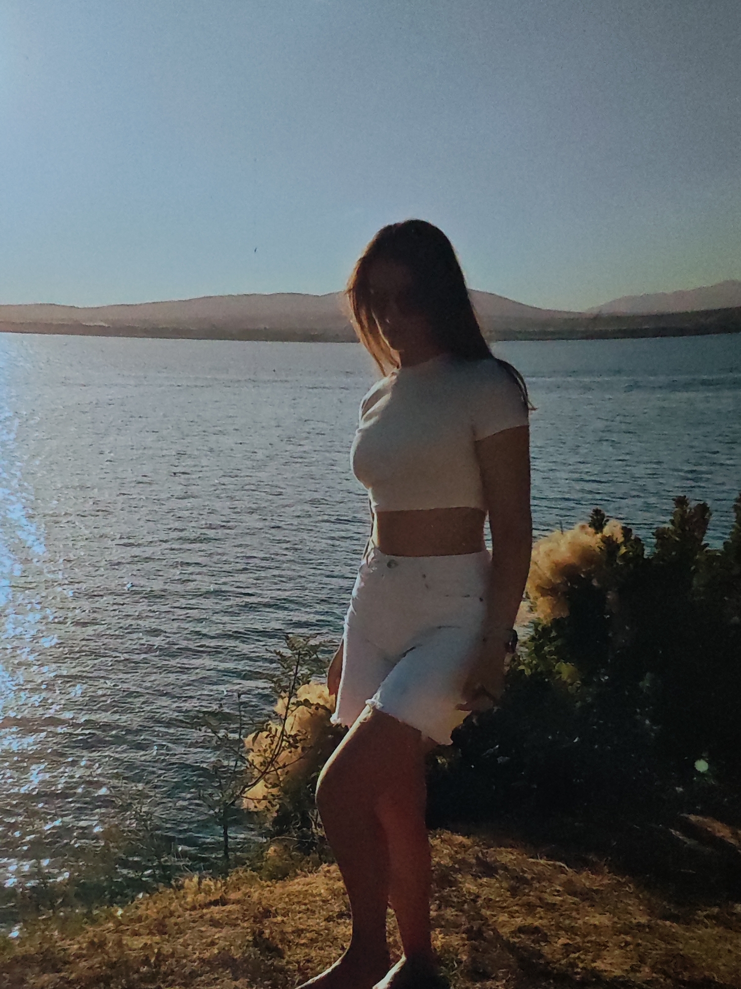 A young woman stands on a grassy embankment overlooking a large body of water. She is wearing a white crop top and white shorts. The water is calm and reflects the clear blue sky. In the distance, hills are visible across the water. There is vegetation in the foreground and on the embankment. The lighting suggests it is either early morning or late afternoon, with the sun casting a warm glow on the scene.