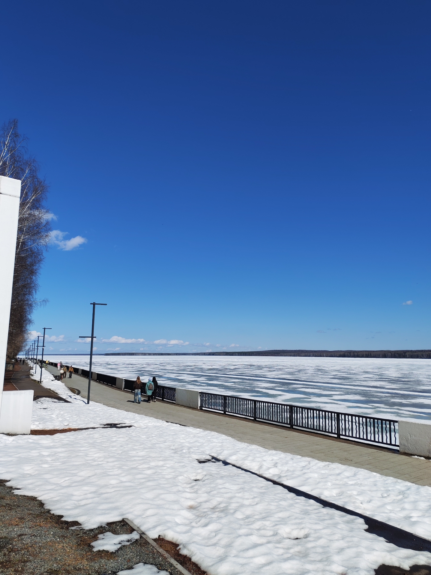 The image depicts a waterfront scene in Chaikovsky, Russia, characterized by a clear, blue sky and a landscape transitioning from winter to spring. A partially frozen body of water, likely the Kama River or a reservoir, stretches across the midground, showing large sheets of ice with open water in between.
Along the waterfront, a paved promenade is visible, flanked by a black metal railing that separates it from the water. The promenade shows patches of melting snow, indicating the onset of warmer weather. A line of evenly spaced, modern lampposts runs along the promenade.
Several people are walking on the promenade, dressed in casual clothing appropriate for the weather. To the left of the promenade, there is a small strip of land with more melting snow and some exposed ground. Bare trees stand along the edge of the land, suggesting early spring. The scene exudes a sense of quiet activity.