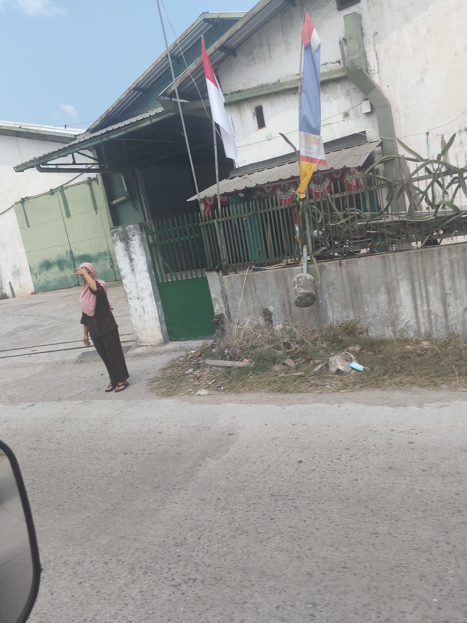 The image depicts an outdoor scene in what appears to be an industrial or commercial area. A woman wearing a pink hijab and dark clothing is standing on a paved surface, looking upwards and shielding her eyes with her hand. Behind her, a building with a green roof and white walls is visible. Two flags are flying from poles in front of the building: one is the Indonesian flag (red and white), and the other is a tricolor flag with blue, white, red, and yellow sections. A green metal fence runs along the front of the building, and beyond it, some dry vegetation and debris are present. The ground in the foreground is asphalt, with a partial view of a vehicle's side mirror on the left. The sky is clear and pale blue. Based on the Indonesian flag and the likely context of such imagery, the location could be Bandar Lampung, Indonesia, a city known for its industrial and commercial activities.