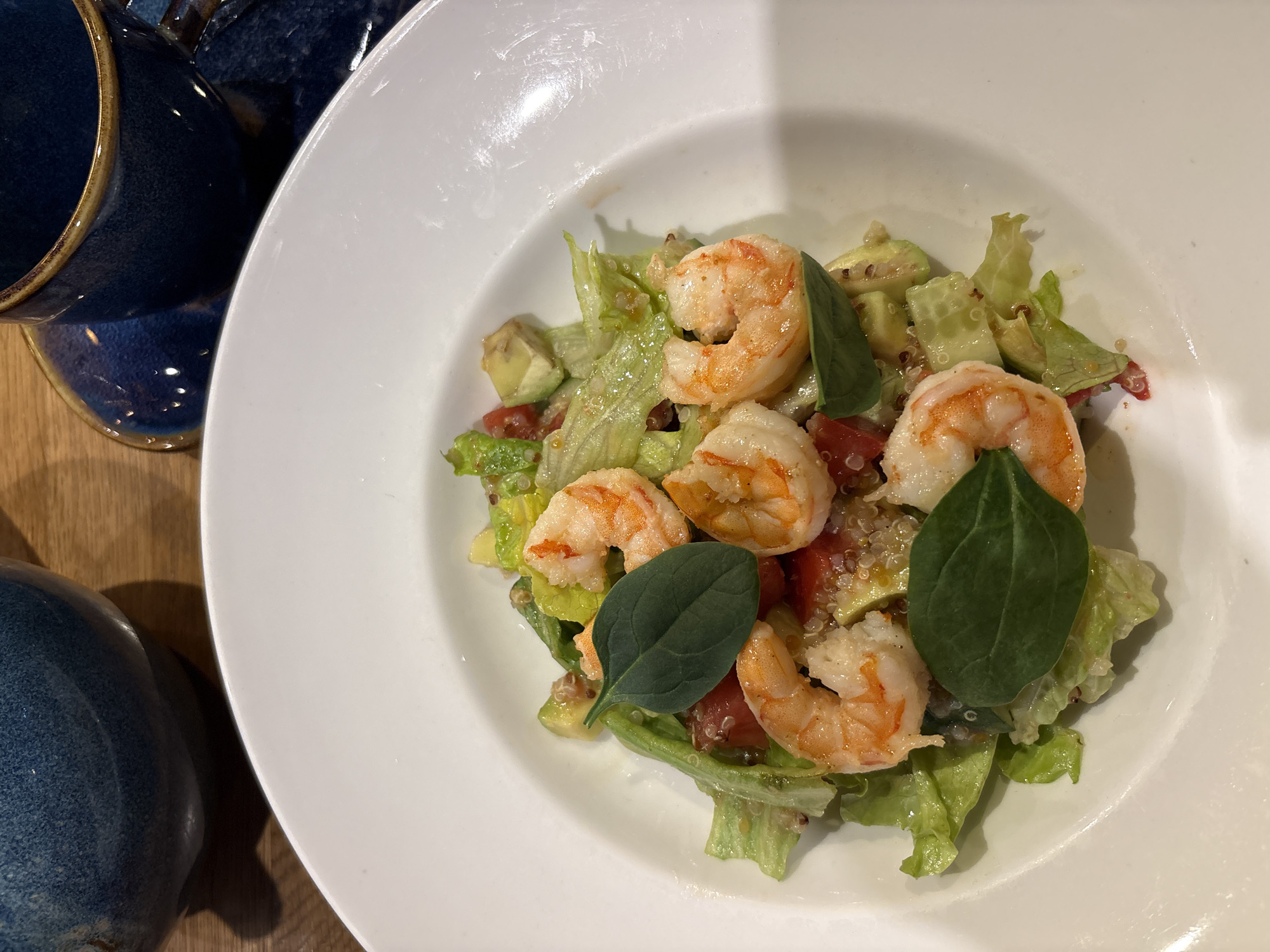 The image depicts a salad with shrimp, lettuce, tomato, avocado, quinoa, and basil, presented on a white plate. A blurred blue ceramic dish is positioned to the left of the plate. The setting appears to be indoors, possibly a restaurant or dining area, with a wooden table surface visible. Kazan, Russia is not explicitly verifiable in the image, but possible based on restaurant data.