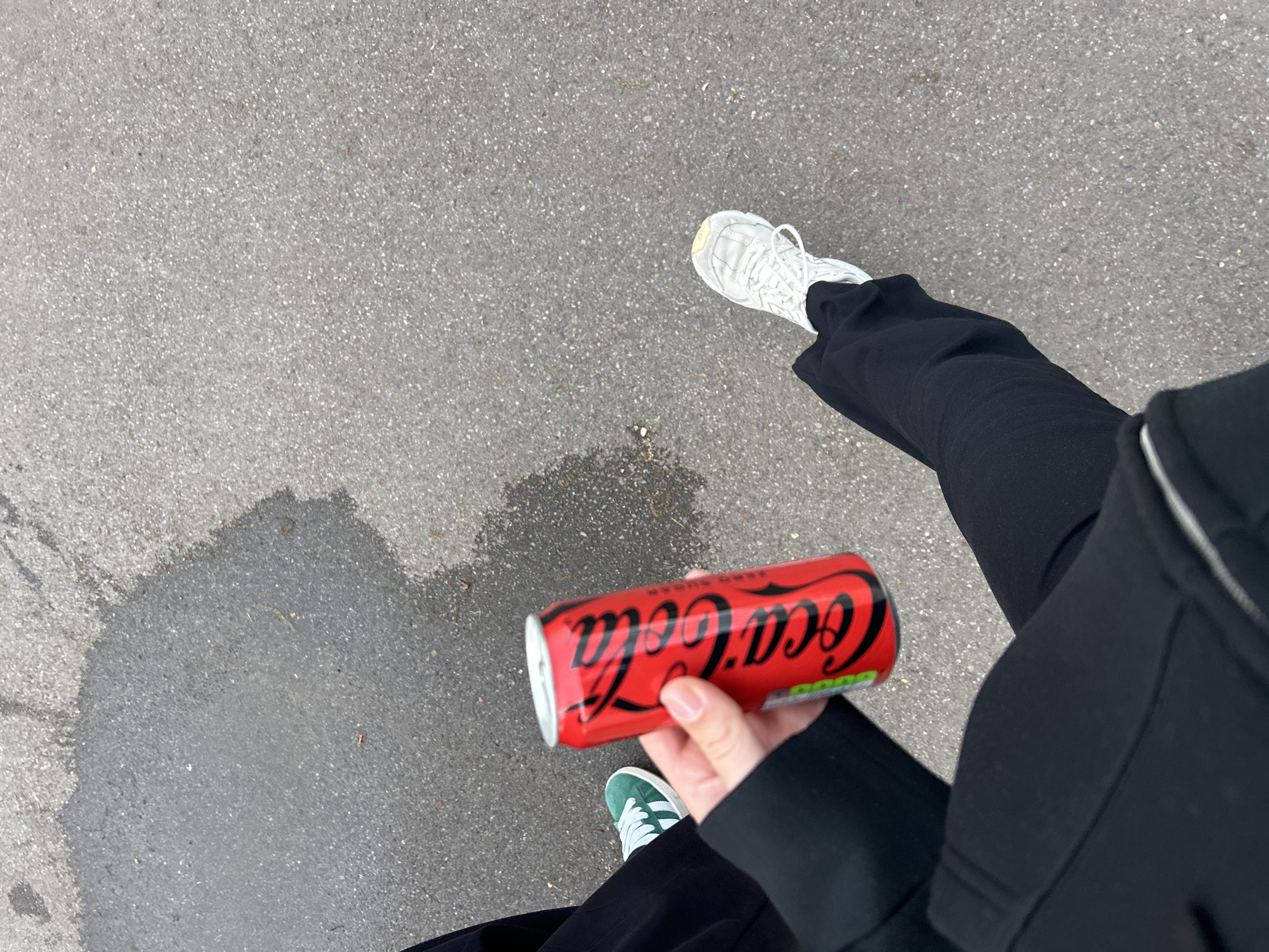 A first-person perspective photograph shows a person holding a red Coca-Cola can. The person is wearing black pants and a black jacket. The person is also wearing one white sneaker and one green sneaker with white stripes. The person is standing on a gray asphalt surface. The person took this photo in Noisiel, France.