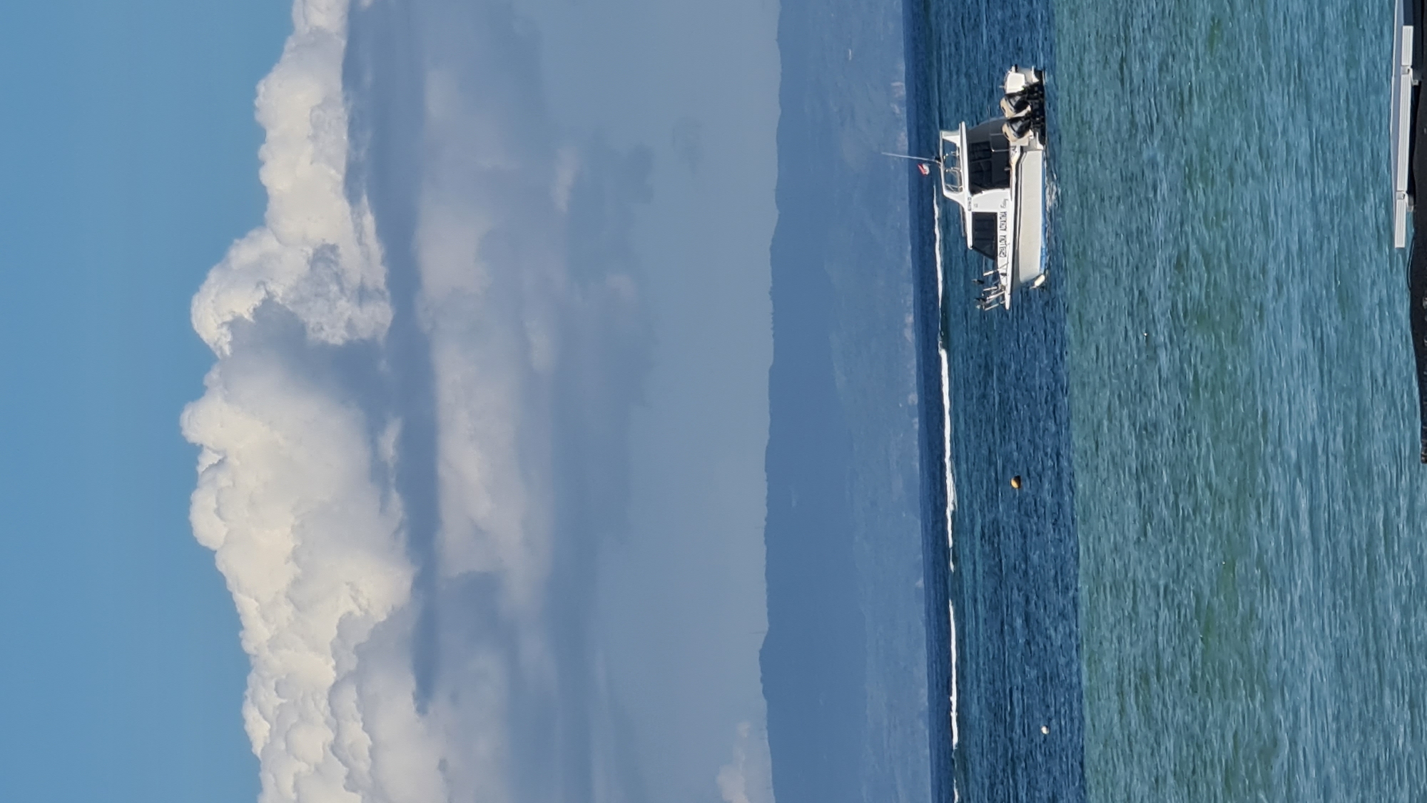 A white motorboat with twin outboard engines is anchored in the ocean, with a mountainous coastline in the background. The boat has the text "GENULOKA ADYATHA" written on its side. Two buoys are visible in the water near the boat. The sky is filled with large, puffy white clouds against a blue sky. The ocean water transitions from a deep blue to a lighter turquoise. The scene appears to be taken from a shoreline or a floating structure. The location is likely the coast of Denpasar, Indonesia, given the mountainous background typical of Bali and the presence of a boat that could be used for local fishing or tourism.