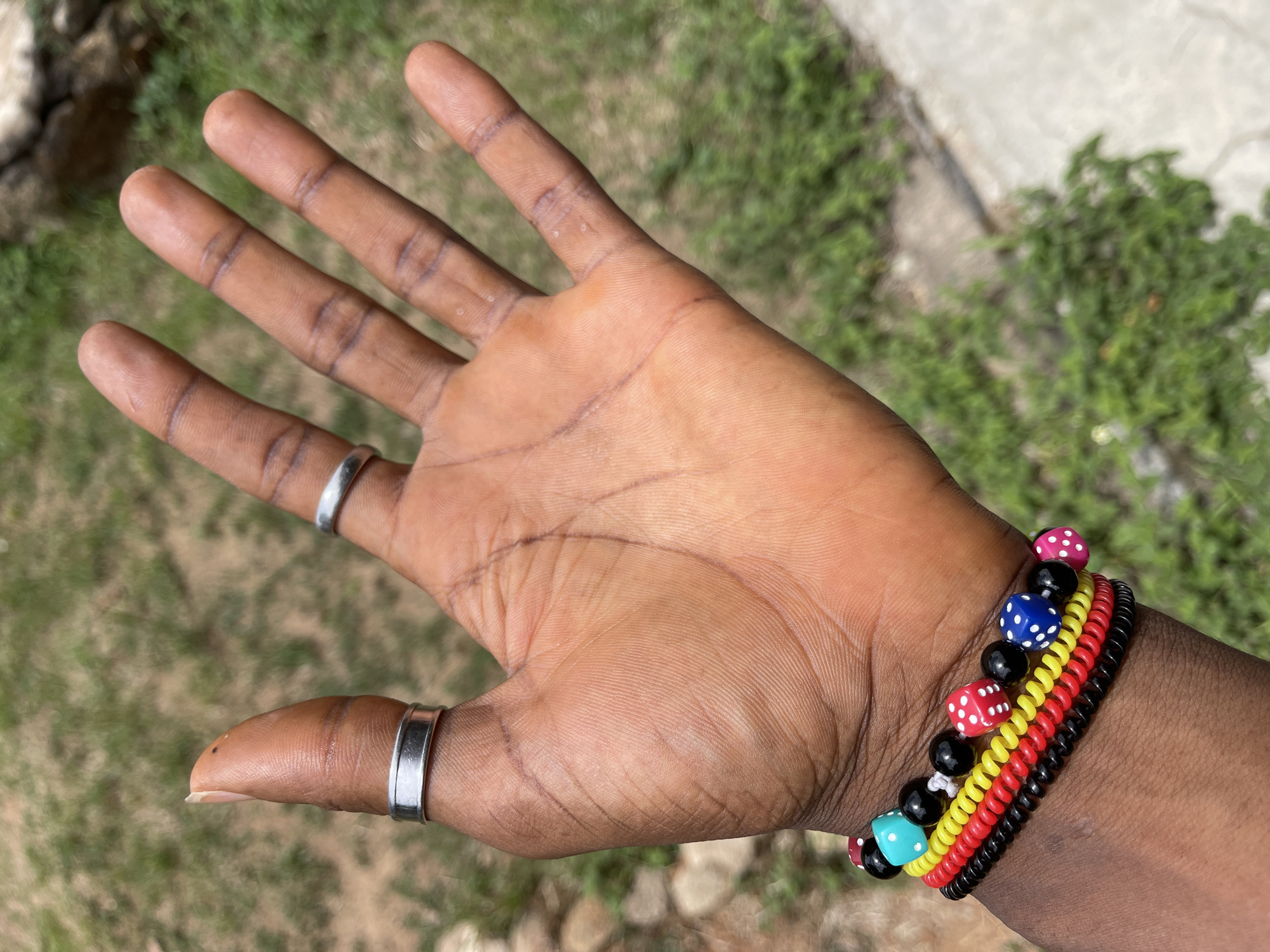 An open dark-skinned hand is centered in the image, with the palm facing the viewer. The hand has two silver rings, one on the thumb and one on the index finger. Several bracelets adorn the wrist, including beaded bracelets with dice-shaped beads and colorful spiral bracelets. The background is blurred, showing green vegetation and a light-colored surface, possibly stone or concrete. There is no clear indication of a city or specific location such as Jos, Nigeria, present in the image.