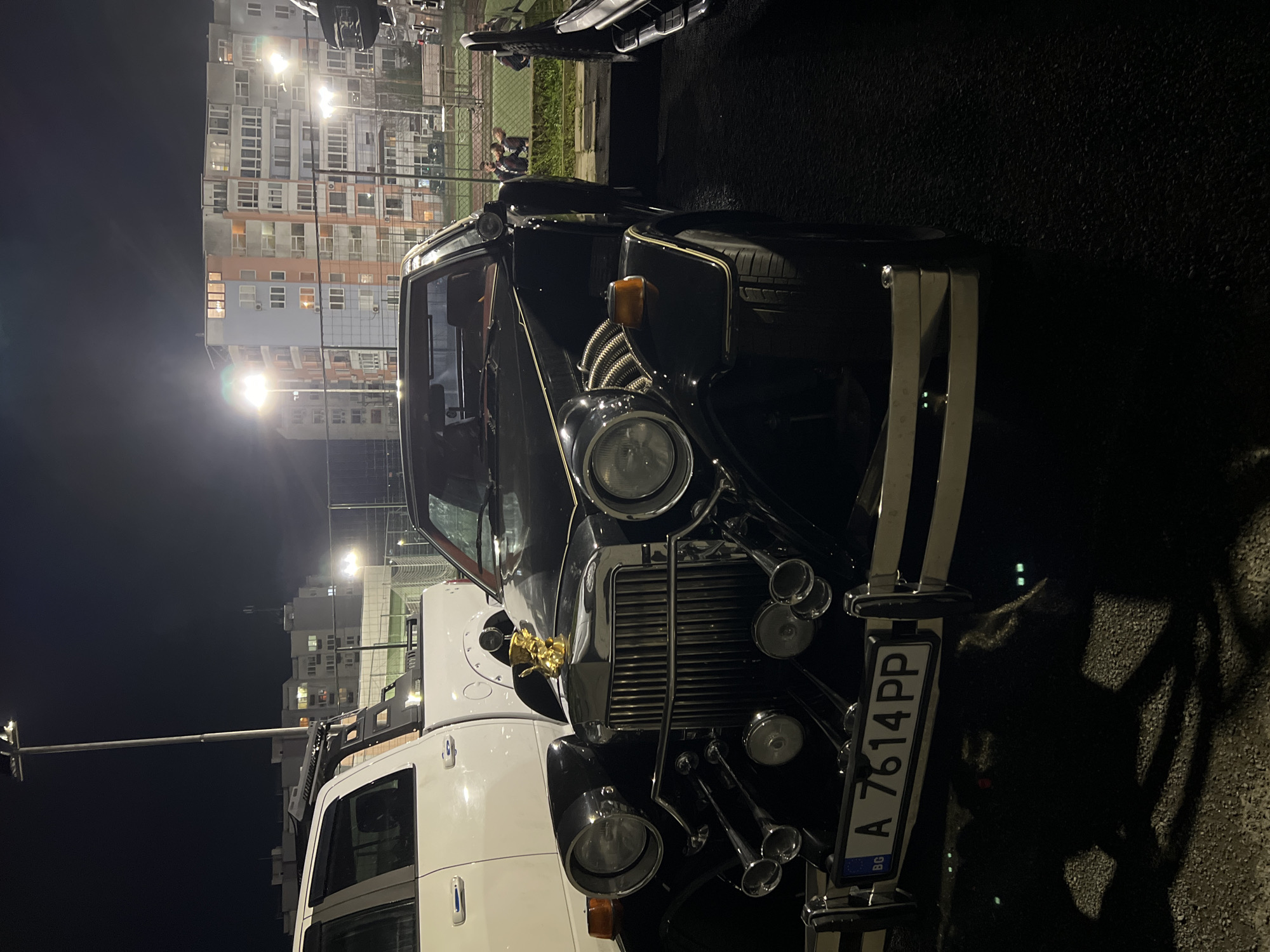 The image shows the front of a black vintage-style car with a Bulgarian license plate reading "A 7614 PP." The car has a prominent grill, multiple horn-like fixtures, and a golden hood ornament. A white vehicle is partially visible behind the car. The background includes a brightly lit apartment building and streetlights, suggesting an urban setting at night. Based on the license plate, the location is in Bulgaria. While pinpointing the exact location in Burgas is impossible without more information, the presence of a large apartment building and streetlights suggests a residential area or a parking lot near such an area. In the background, near a fence, three people can be seen.
