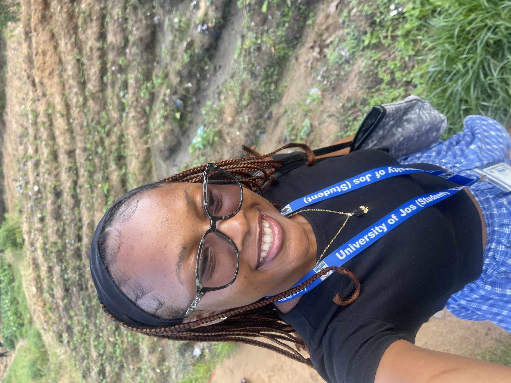 A woman is taking a selfie outdoors in a rural area, with rows of cultivated land visible in the background. She is wearing a black t-shirt, a black headband, eyeglasses, and a blue lanyard with the text "University of Jos (Student)". She is also wearing a blue plaid skirt and a gold necklace with a dark pendant. Her hair is styled in braids. The background shows a field with some vegetation and dirt paths. There are indications of some debris scattered in the field. The lighting suggests it is daytime, likely overcast or late afternoon. The overall impression is of a student in an academic or community setting, possibly engaged in fieldwork or a campus-related activity given the lanyard. The location is identified as Rafin Par, Nigeria.