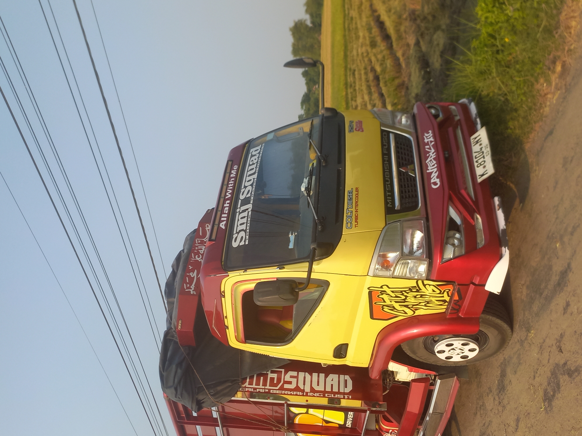 A yellow and red Mitsubishi Fuso truck is parked on a dirt road next to a harvested rice field in Tamping Mojo, Indonesia. The truck has several decals and text on it, including "Allah With Me," "Mjn Squad," and "DRIVER." The license plate reads "K 8304 NY." The sky is clear and blue. The scene appears to be a rural area.