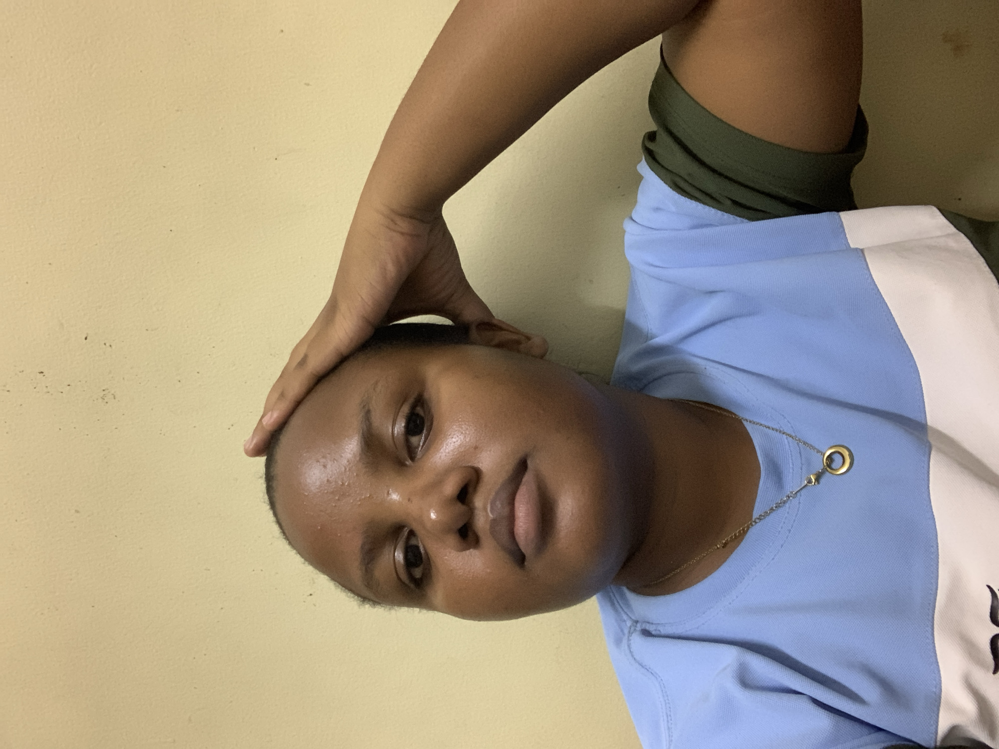 A young woman is depicted indoors, likely in a private residence, given the plain yellow wall in the background. Her arm is raised, hand placed atop her head in a casual gesture. She is wearing a blue, white, and possibly green top, and a gold necklace is visible. The lighting is soft and indirect. The location in the city of Dar es-Salaam, Tanzania cannot be verified from the image.