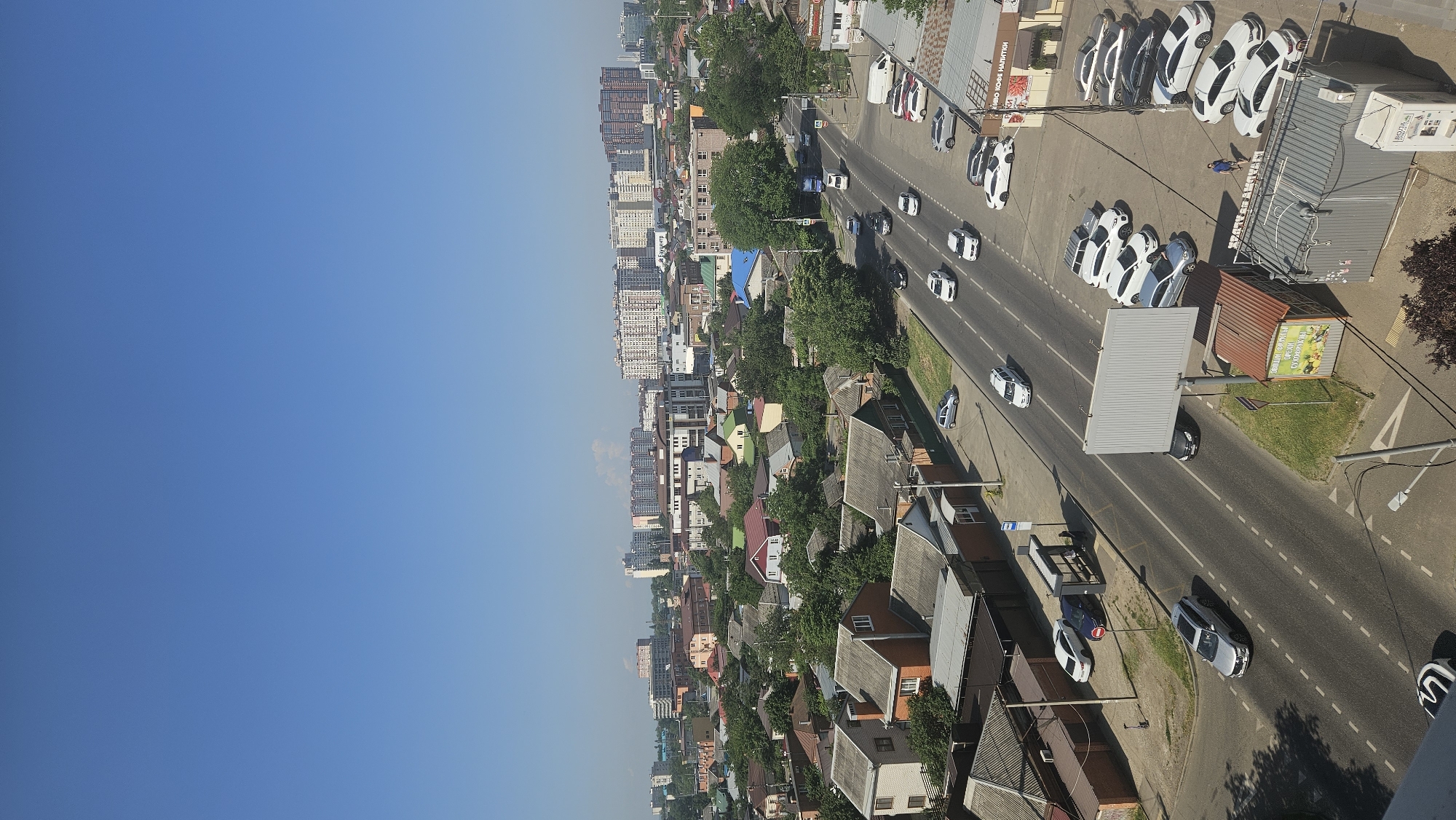 The image shows an aerial view of a city street with buildings, trees, and vehicles. The sky is clear and blue. The street is asphalt with white dashed lines indicating lanes. Several cars are driving on the street and parked on the side. There are houses with grey and red roofs, and some larger apartment buildings in the background. A bus stop is visible on the side of the road. A large billboard stands next to the street. There are also some commercial buildings with signs. A single person is walking on the sidewalk near the parked cars. The overall scene depicts a typical urban environment on a sunny day.