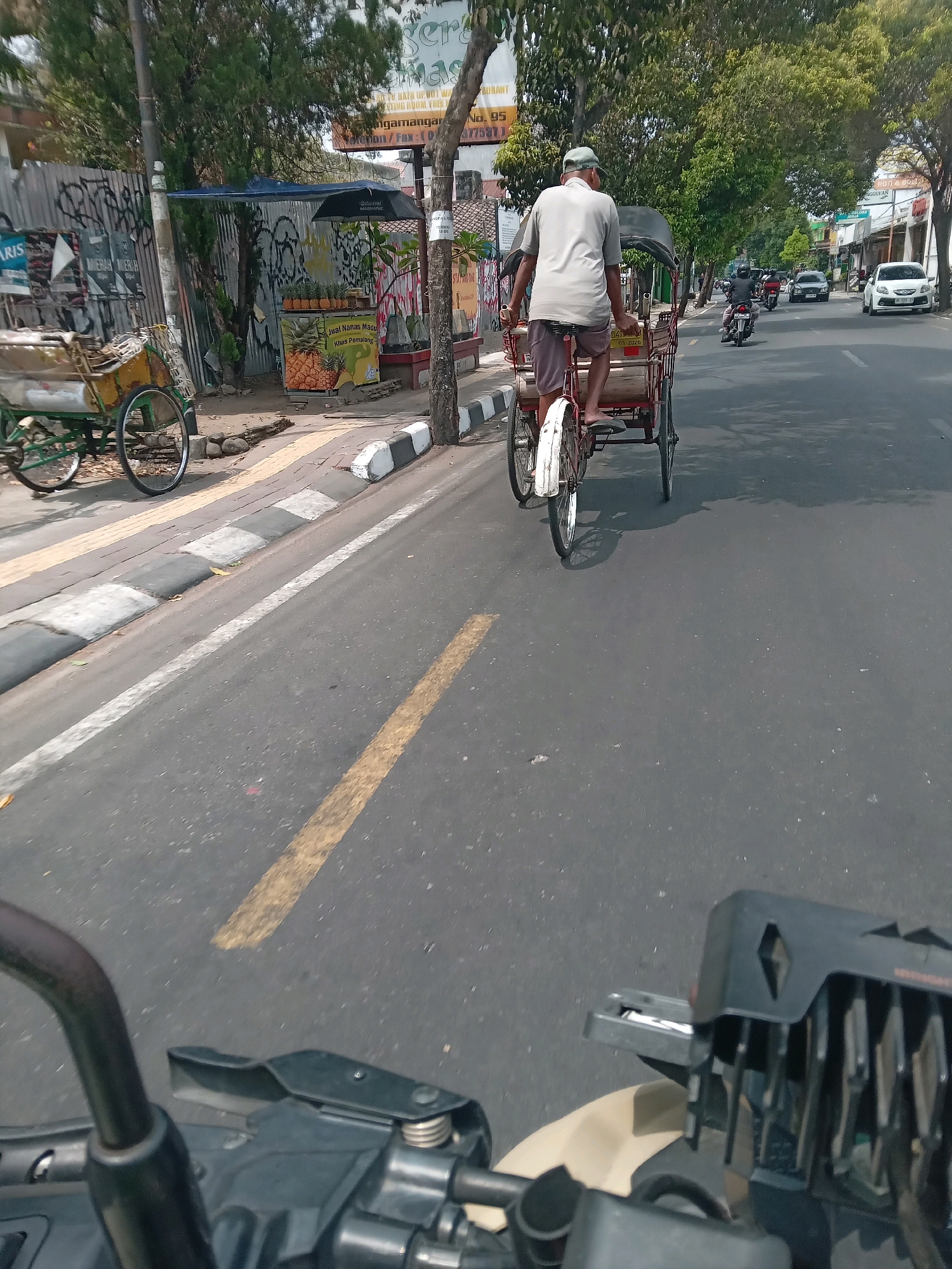 The image captures a street scene in Yogyakarta, Indonesia, likely on a sunny day. In the foreground, the handlebars and dashboard of a motorcycle are visible, suggesting the photo was taken from a rider's perspective. The motorcycle is on an asphalt road marked with yellow and white lines.
Further ahead, a traditional becak (pedicab) is being ridden by a man wearing a light-colored cap and t-shirt, and shorts. The becak has a red frame and two wheels at the back. Beside the road, a cart with green framing, possibly another form of transport or stall, is parked.
The background shows a sidewalk with a black and white striped curb. A fruit stand selling pineapples is visible, along with a blue umbrella providing shade. Trees line the street, and buildings with shopfronts can be seen in the distance. Other vehicles, including motorcycles and cars, are also present on the road, indicating typical city traffic. Graffiti is visible on a corrugated metal fence on the left side of the image. The overall atmosphere suggests an active urban environment.