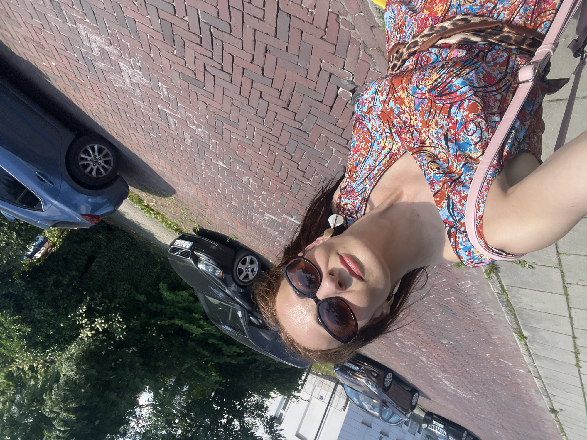 A woman wearing a colorful dress and sunglasses takes a selfie outdoors on a brick street. She has a pink purse slung over her shoulder. In the background, several cars are parked along the street and a line of trees borders the sidewalk. The environment appears to be a sunny day in a residential or commercial area of a city.