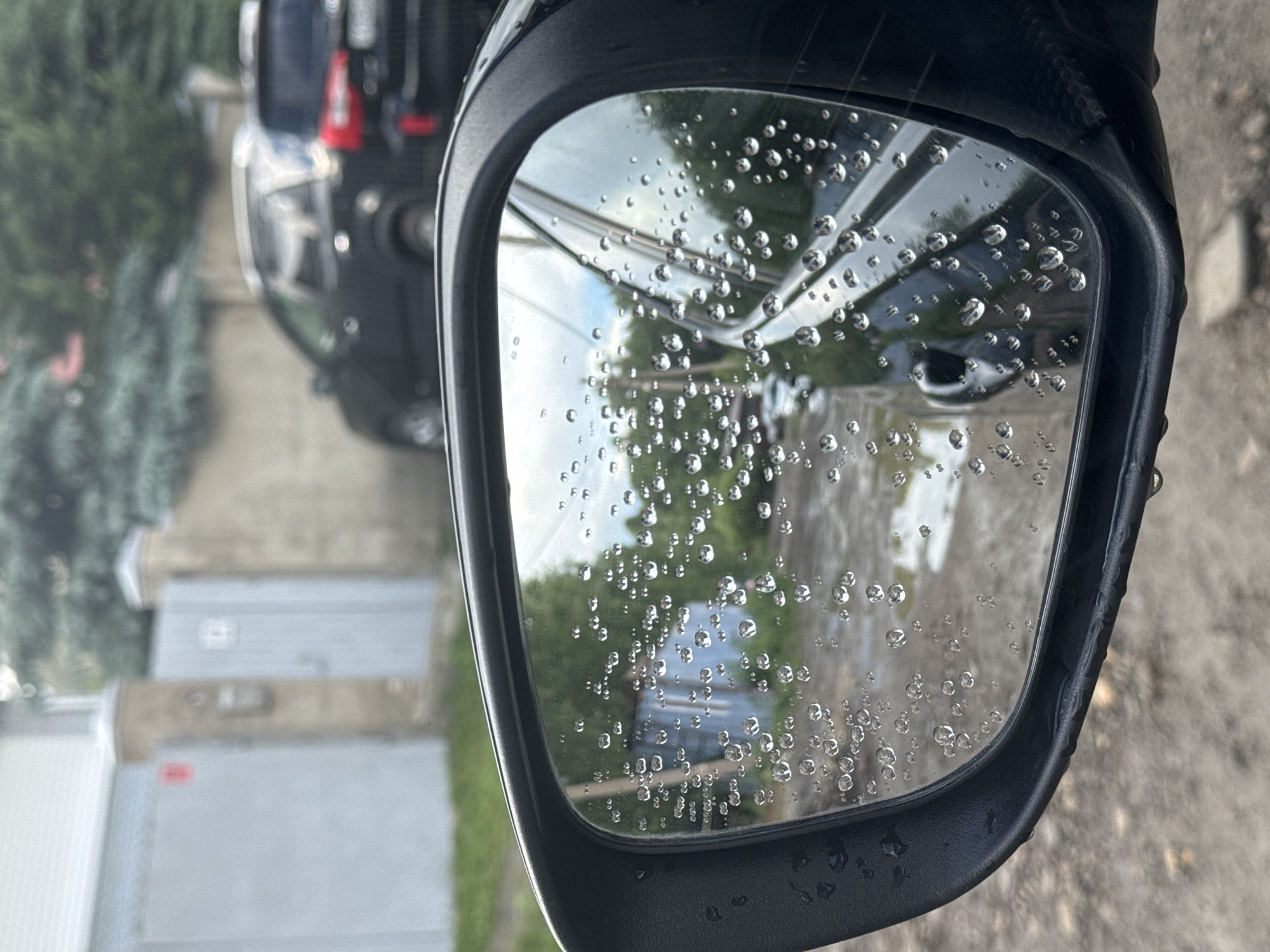 The image is a close-up of a car's side-view mirror, which is covered in raindrops. The mirror reflects a blurry scene of a street with puddles, trees, and buildings. In the background, there are two dark-colored SUVs parked on the side of the street. The overall impression is that of a rainy day in a residential area. The location is identified as Кусимовского рудника, Russia.