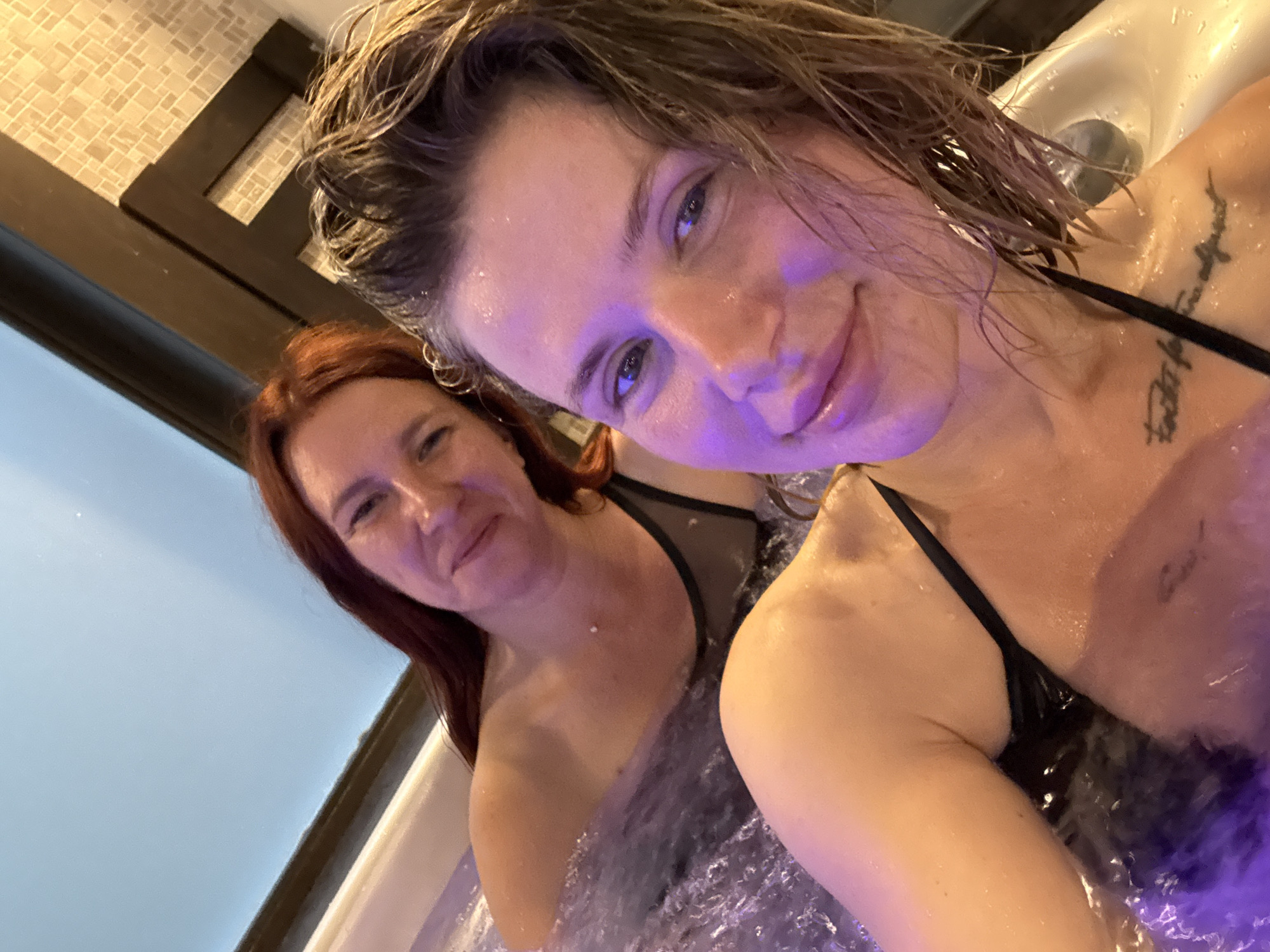 Two individuals are in a hot tub filled with bubbly water. The person in the foreground, who is closer to the camera, has wet blonde hair and is wearing a black swimsuit. A tattoo is visible on their collarbone. The person in the background has long red hair and is also wearing a black swimsuit. They are smiling at the camera. The hot tub appears to be indoors, with a tiled wall visible in the background and a light blue ceiling. The location is not specified as Urlu-Aspak, Russia, as that information is not present in the image.