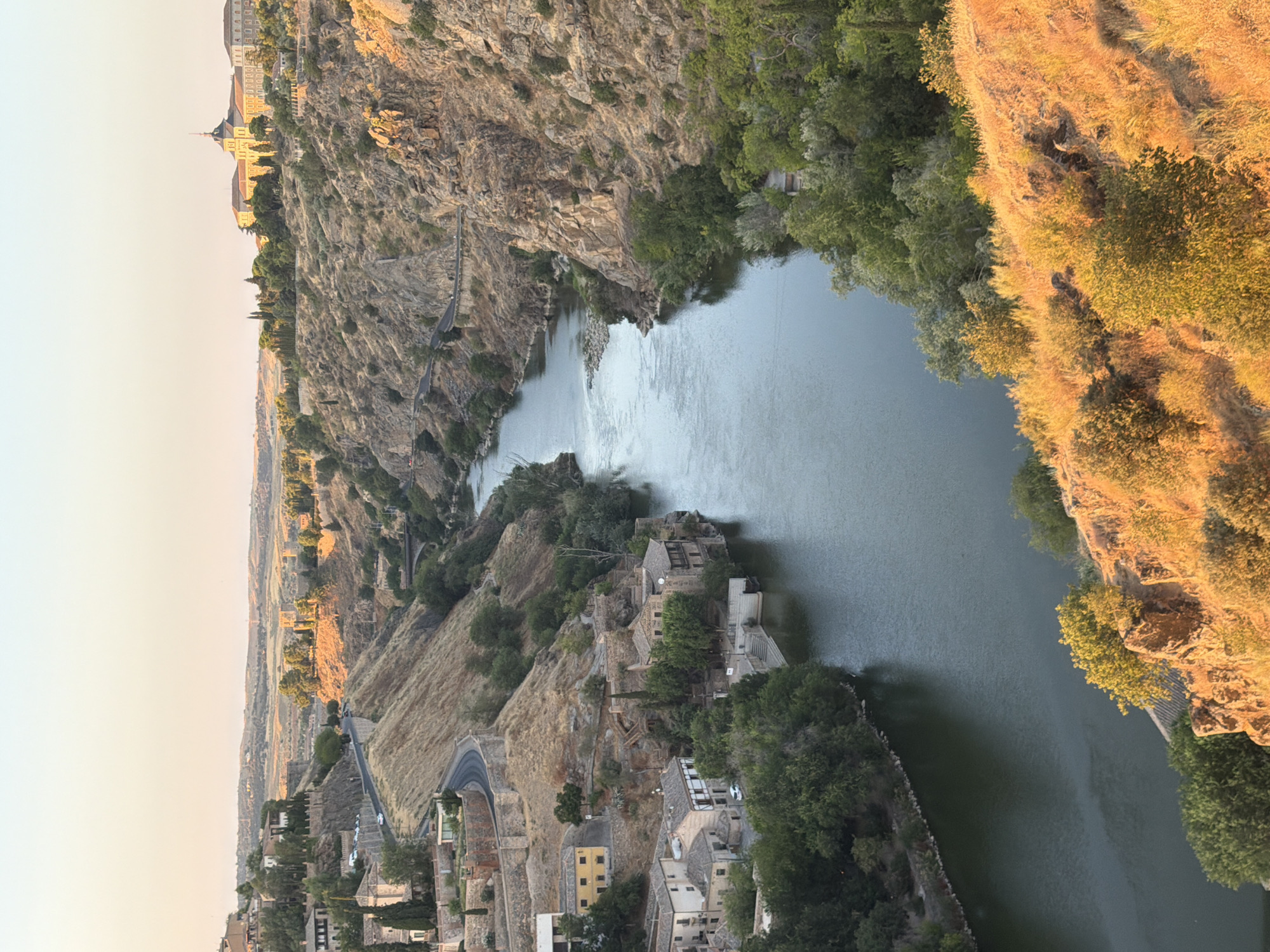 The image depicts a panoramic view of Toledo, Spain, showcasing the Tagus River winding through a rocky gorge. On the left bank, a cluster of ancient stone buildings with red-tiled roofs clings to the hillside, interspersed with lush green trees. A winding road is visible above the buildings, with a few cars parked along its edge. The right bank is characterized by steep, rocky cliffs, also sparsely vegetated with trees and shrubs. In the distance, on the elevated plateau overlooking the river and gorge, a large, imposing building with a central dome and a lighter colored facade stands prominently, possibly a historical monument or religious institution. The sky above is a pale, clear gradient of orange and yellow, indicating either sunrise or sunset. The overall impression is one of historical grandeur and natural beauty, with the river serving as a central artery through the dramatic landscape.