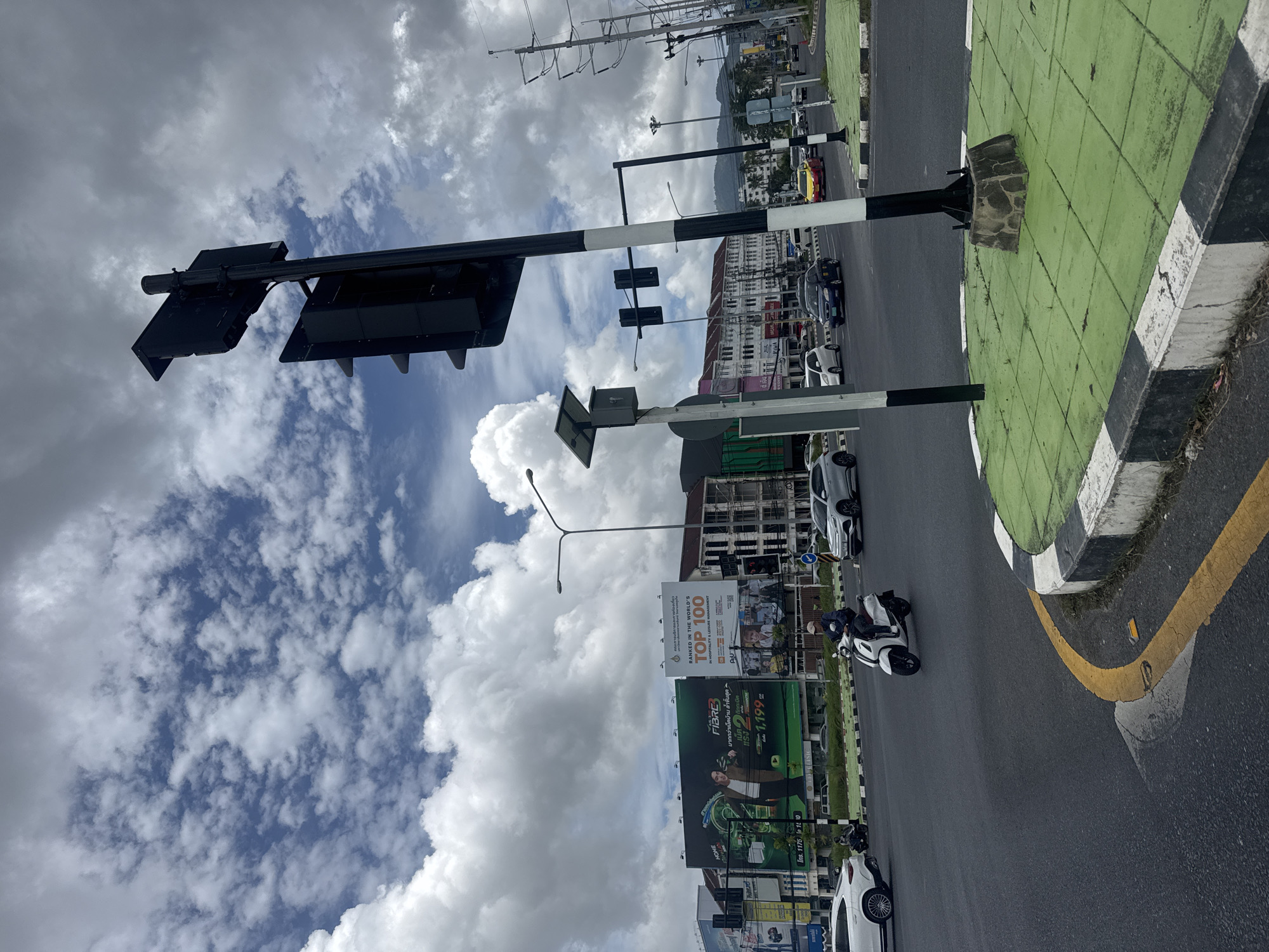 The image depicts a street intersection in Ratsada, Thailand, under a cloudy sky. A traffic light stands prominently in the foreground, with a dark pole and multiple signal heads. The intersection is paved with asphalt, and a circular green island with a black and white striped curb is visible. A white scooter with a rider, dressed in a dark jacket and a helmet, is in motion at the intersection. Several cars, including a white SUV, a blue sedan, and a yellow taxi, are also present on the road. Large billboards are mounted on buildings lining the street. One billboard features an advertisement with a man and the text "HOME FIBRE". Another displays "RANKED IN THE WORLD'S TOP 100 IN HOSPITALITY & LEISURE MANAGEMENT". Power lines and poles are visible in the background, along with buildings and distant hills. The overall scene suggests an active urban environment.
