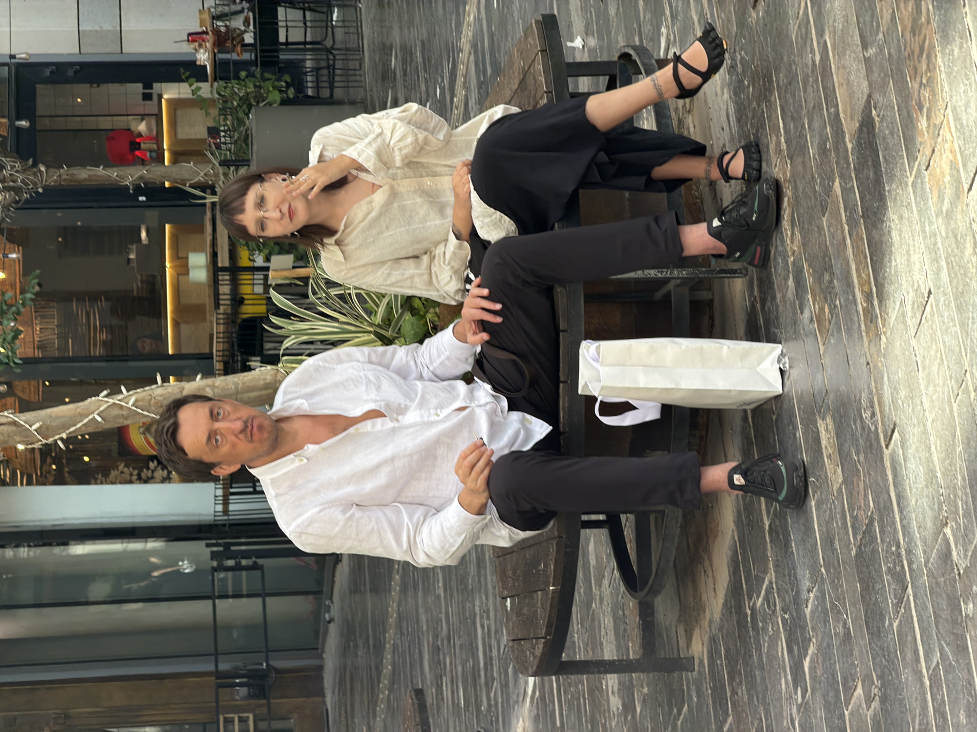 Two people are sitting on a park bench. The woman is wearing a light-colored button-up shirt and black pants with black sandals. The man is wearing a white button-up shirt and black pants with black sneakers. They appear to be seated near a building in an urban environment, possibly a cafe or storefront, located in Tel Aviv, Israel. The seating area is paved with patterned stones, and there is a white shopping bag on the bench next to the man.