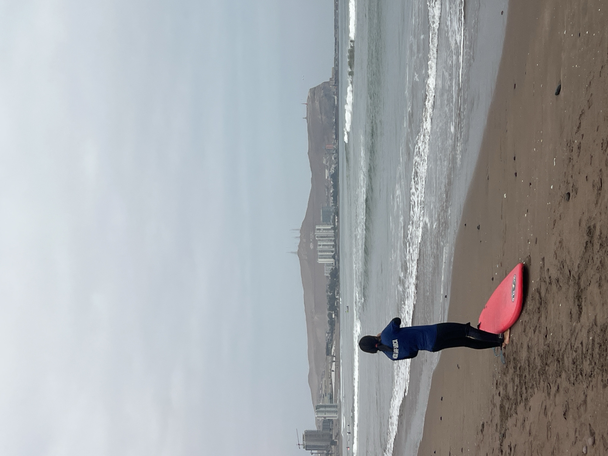 A person stands on a sandy beach next to a red surfboard. The person is wearing dark clothing, likely a wetsuit, and appears to be looking at the ocean. Waves are visible in the background. The cityscape of Arica, Chile, is visible in the background, with the iconic "Morro de Arica" (Arica cape) standing prominently. The sky is overcast.