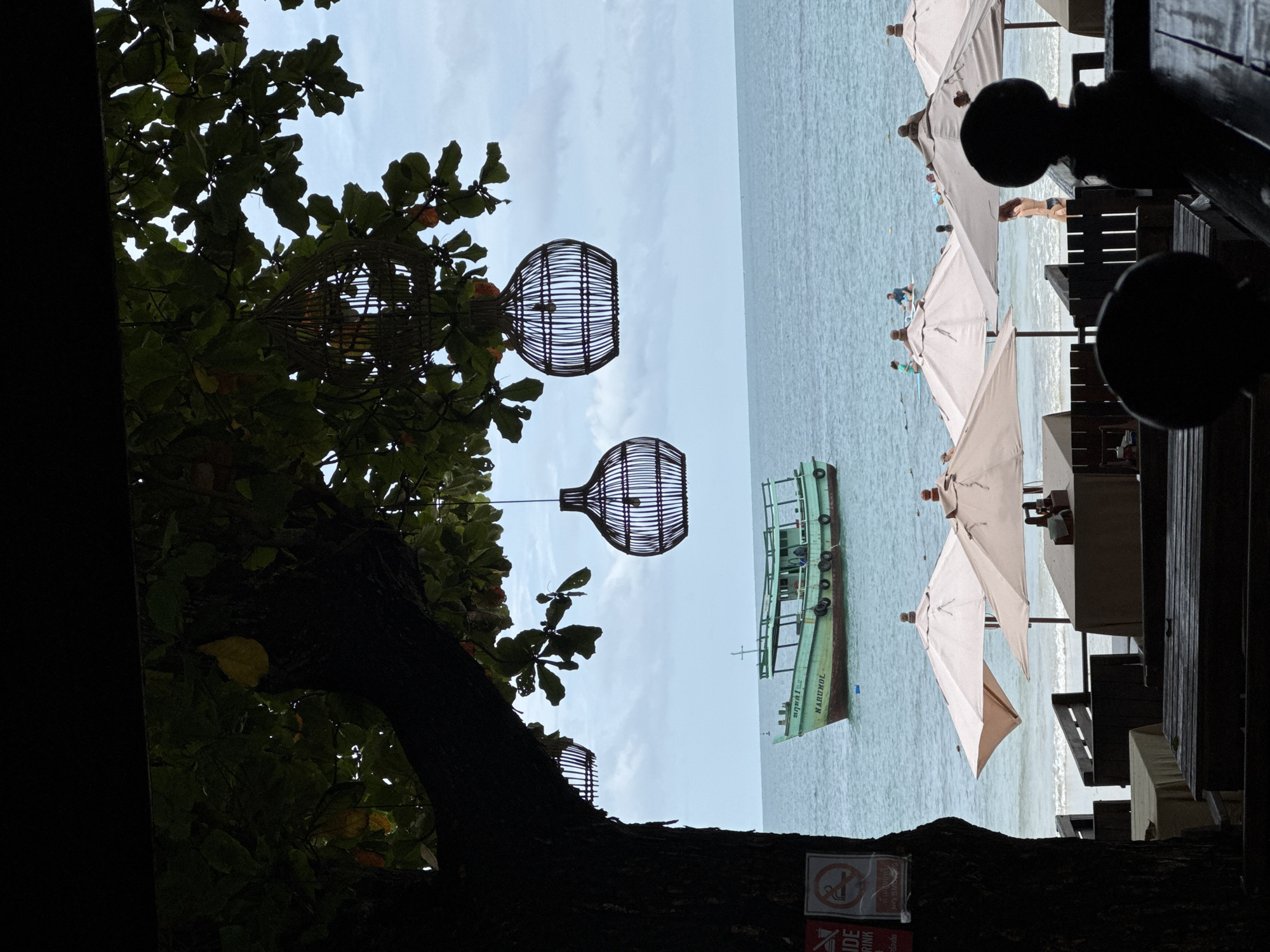 The image depicts a view of Ao Phai beach in Koh Samet, Thailand, looking out towards the ocean. In the foreground, silhouetted against the brighter background, are tables and chairs set up at what appears to be a beachside restaurant or seating area. Wooden structures and umbrellas are visible, with some tables covered in tan tablecloths. To the left, a large tree trunk with dark foliage partially obscures the view, and two circular wicker lanterns hang from its branches. A small sign with a no-smoking symbol is affixed to the tree.
In the middle ground, the calm ocean stretches towards the horizon under a cloudy sky. A light green boat with a cabin and a single mast is anchored offshore, with the name "NARUMOL" visible on its side. Further out, several people are in the water, some on paddleboards and others swimming. Beach umbrellas of various sizes are set up along the shoreline, and faint figures of people can be seen on the beach. The overall scene conveys a relaxed, tropical atmosphere.