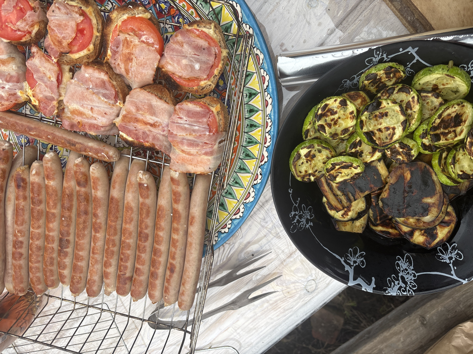 The image depicts a meal likely prepared outdoors. A colorful plate holds toasted bread topped with tomato and bacon, alongside a grill basket of sausages. A black plate to the right contains grilled zucchini and eggplant. The scene is set on a wooden table or surface. The location, Chaikovsky, Russia, is not discernible from the image content alone.