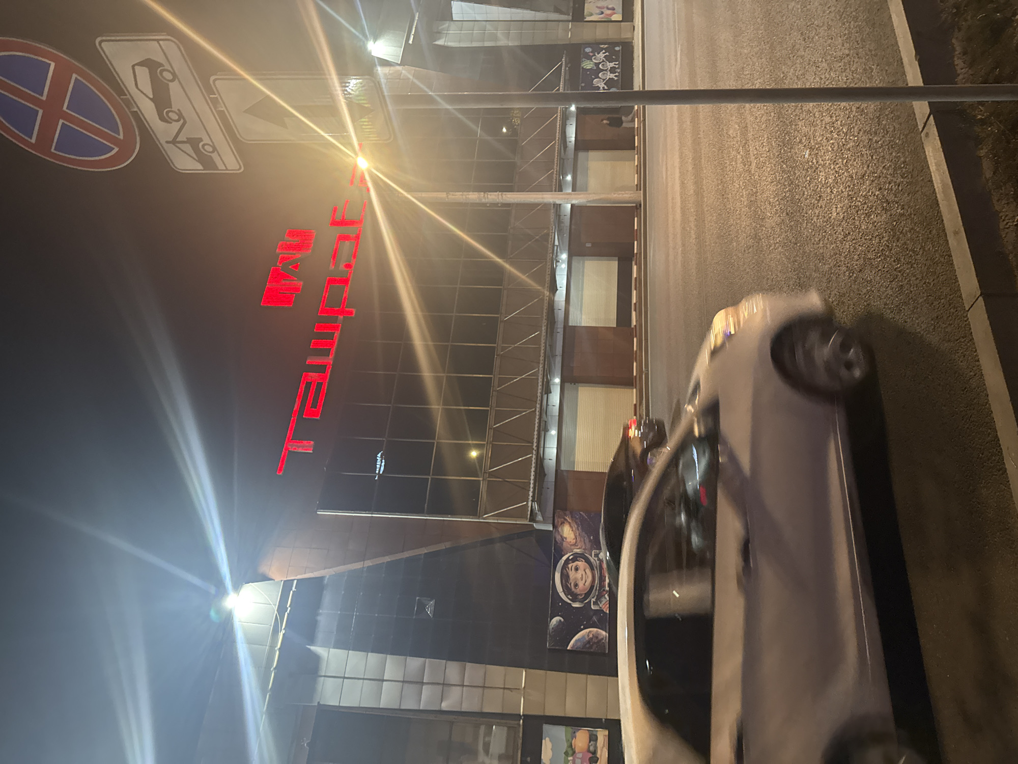 The image shows a street scene at night in Bishkek, Kyrgyzstan. A white car is moving from left to right in the foreground. In the background, there is a modern building with red neon lights spelling out "TAZAPAK". The building has large windows and a dark facade, with some signs visible on the lower level. There are also street signs, including a "no parking" sign and a tow-away sign, mounted on a pole. The street is paved and illuminated by streetlights, creating lens flares in the image.