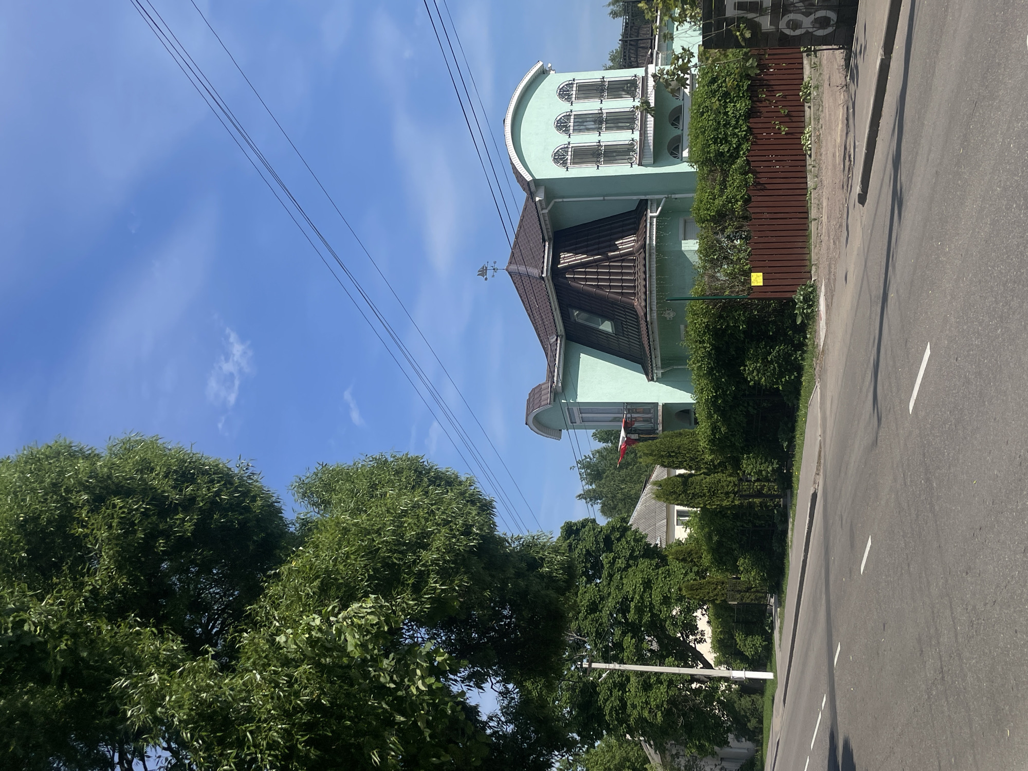The image displays a street scene in Priozersk, Russia, under clear daylight. The foreground features a paved asphalt road with white dashed lane markings. A concrete curb separates the road from a dirt and grass verge on the right, which precedes a sidewalk. Along the property line, a dark brown wooden picket fence is visible, partially obscured by dense green hedges. A small yellow rectangular sign is affixed to a utility pole positioned behind the fence on the left side of the property. To the far right, a concrete structure, possibly a gate pillar or wall, bears the white painted numbers "8" and a partial second digit.
In the midground, a prominent two-story residence with light mint green stucco walls and a dark brown tiled roof stands. The roof exhibits a complex structure with several gables and a large, dark brown tiled dormer. A sailboat-shaped weather vane is mounted on the apex of the main roof. The house features multiple arched windows with white frames and decorative black iron grilles on its second story, notably a cluster of three on the right facade. Two balconies are present: a smaller one on the left with a reddish fabric object draped over its railing, and a wider balcony on the right extending across the three arched windows. The house is surrounded by manicured green bushes and slender, conical evergreen trees. To the left of this house and further back, a portion of another residence with light-colored walls and a grey roof is visible, partially obscured by the extensive green canopy of a large deciduous tree. A white utility pole stands on the left side of the road.
The upper section of the image shows a bright blue sky with scattered, wispy white clouds. Several black power lines stretch horizontally across the sky from the left to the right. No individuals or animals are present in the scene. The lighting suggests a sunny day with discernible shadows.