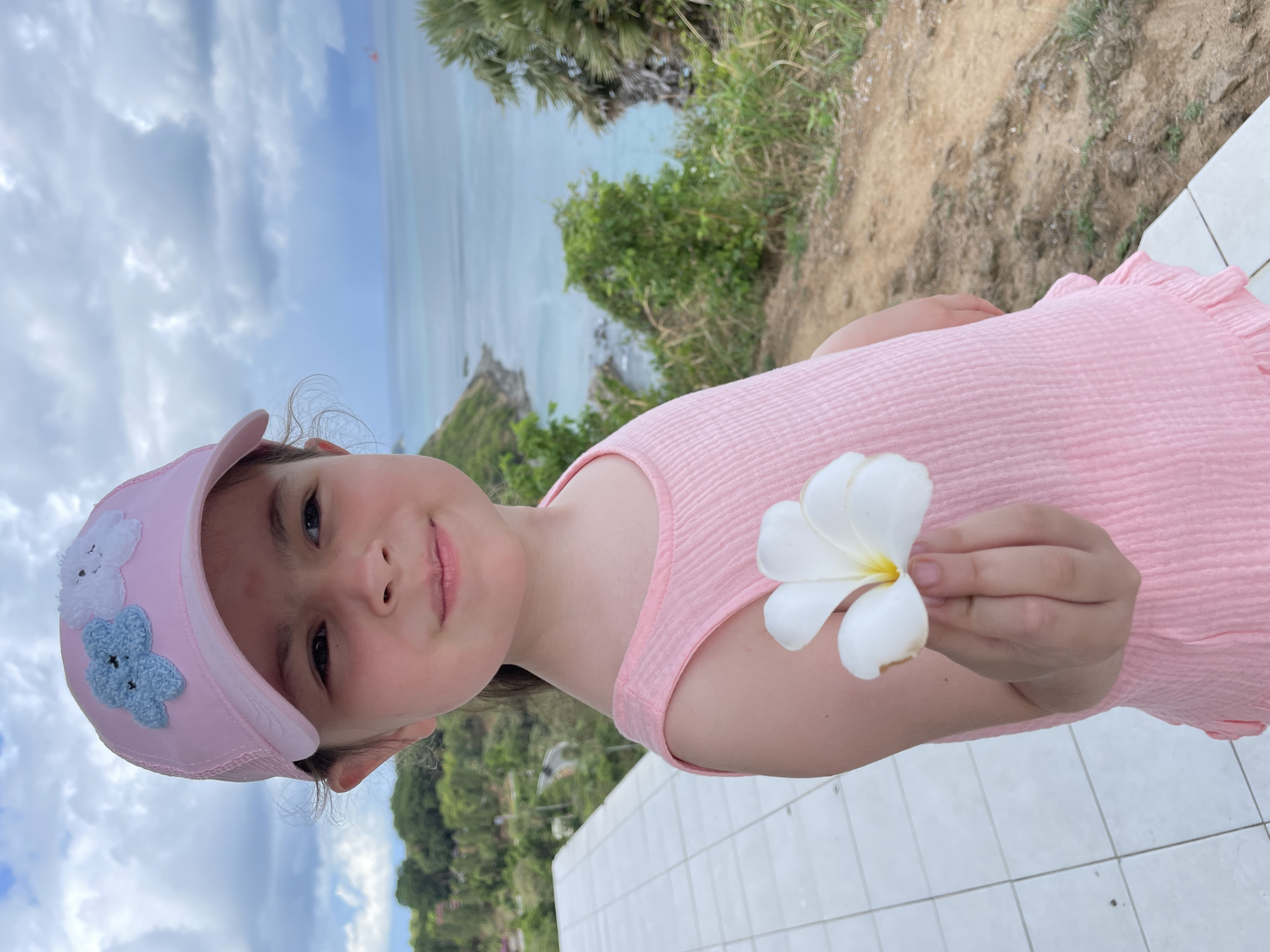 A young girl is holding a white Plumeria flower in her right hand. She is wearing a pink dress and a pink baseball cap adorned with crocheted flowers. The background reveals a scenic landscape including a blue ocean, lush green vegetation, and a cloudy sky. The location is likely near a coastal area in Rawai, Thailand, based on the presence of tropical foliage and the ocean view. The girl is standing next to a tiled surface, possibly a wall or a deck. The image is rotated counter-clockwise.