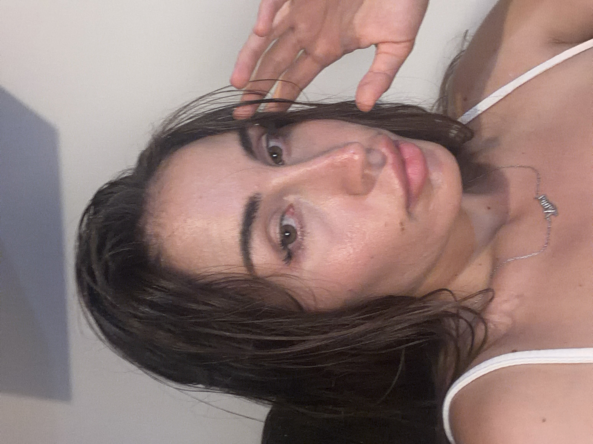 A close-up, shoulder-up, selfie-style portrait shows a young adult woman with dark, wet hair, looking directly at the camera. Her right hand is raised to her forehead, obscuring part of her hair. She is wearing a white tank top and a silver necklace with a pendant. The background is a plain, light-colored wall. The image appears to have been taken indoors. There is no indication of the location within Tersefanou, Cyprus.