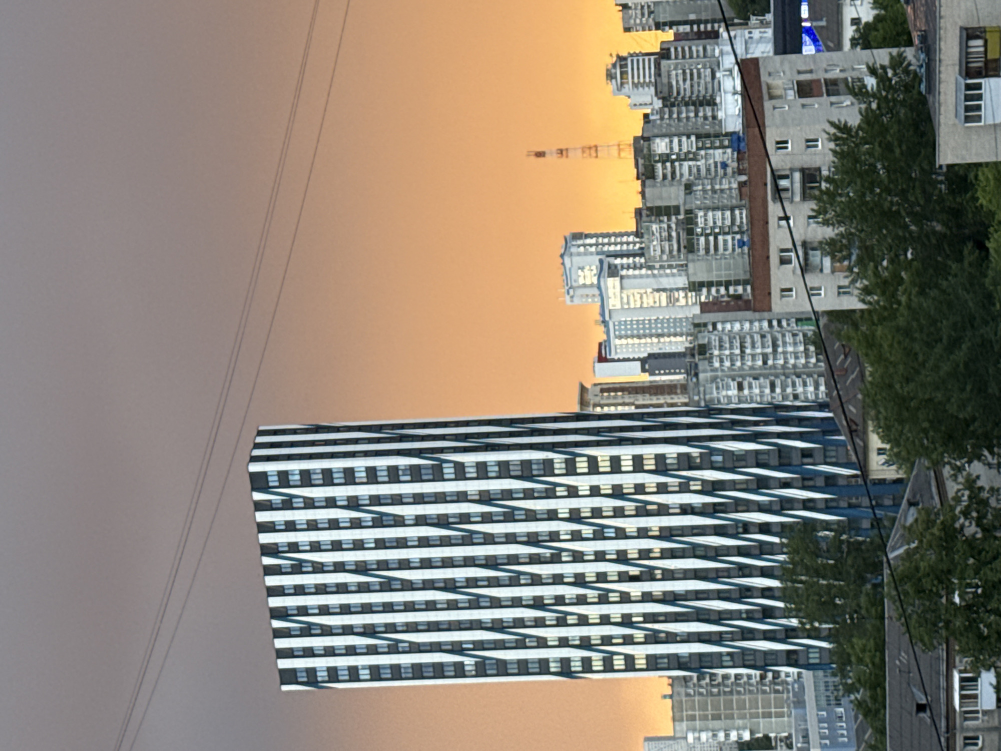 The image shows a view of Yekaterinburg, Russia, at dusk. The sky transitions from a hazy grey to a vibrant orange near the horizon. Prominently featured is a modern building with a striped pattern of windows, contrasting light and dark elements. In the background, traditional Soviet-era apartment blocks are visible, juxtaposed with a construction crane. A high-voltage power line stretches across the upper left of the frame. The composition includes some greenery from trees interspersed among the buildings.