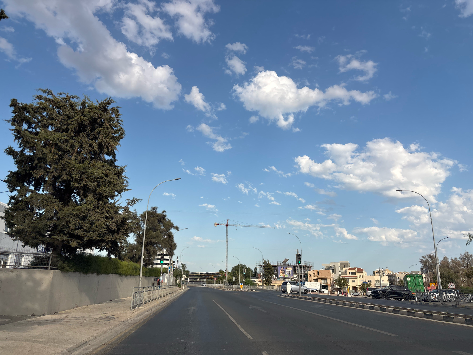The image depicts a view from a moving vehicle on a street in Mesa Geitonia, Cyprus. A wide asphalt road with lane markings dominates the foreground, curving slightly towards the right. On the left side of the road, there is a sidewalk and a grey concrete wall topped with dense green foliage and a large, mature evergreen tree. Further left, a building with a white facade is visible behind a metal fence.
The right side of the road features a sidewalk with a metal railing. Beyond the railing, there is a low barrier with black and white checkered markings. Several cars are parked or driving on this side, including a black SUV, a silver sedan, and a white truck. Buildings of various architectural styles, including residential and commercial properties, line the street. Some buildings appear to be under construction, with a large crane visible in the distance.
Traffic lights with green signals are present at an intersection ahead. The sky is a clear blue with scattered white, fluffy clouds. Streetlights on tall poles are visible along the road. The overall scene suggests a typical urban environment on a clear day, with active construction and traffic.