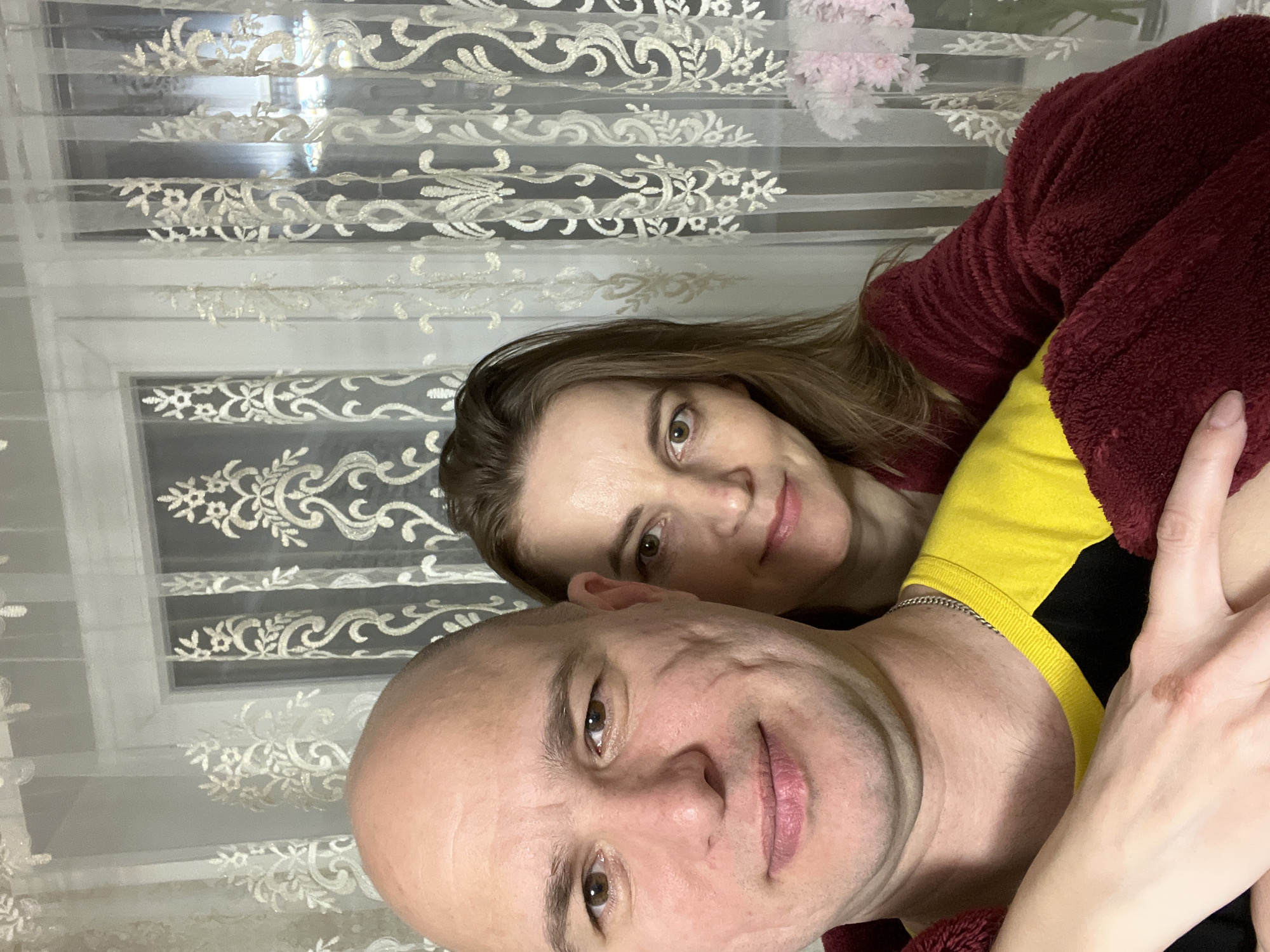 A selfie shows a man and a woman with a window behind them. The man is bald and his face is toward the camera. The woman is looking at the camera, but her face is partially obscured by the man. The man is wearing a shirt that is yellow with a black chest, and a chain around his neck. The woman is wearing a maroon zip-up hoodie. The window behind them has white lace curtains. Location and city are unknown.