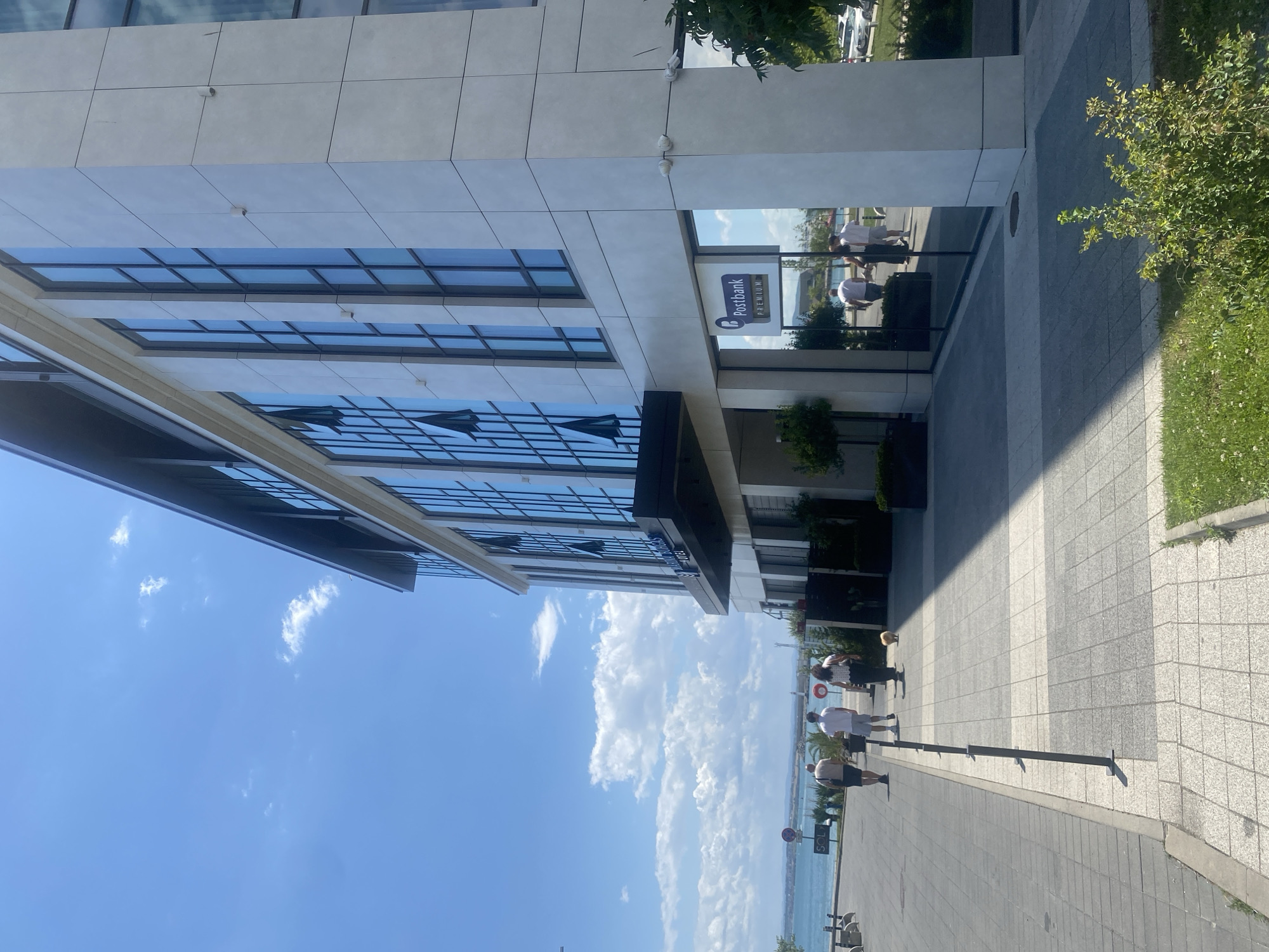 The image shows a modern building with large windows, likely a hotel or office complex, located in Burgas, Bulgaria. The building's facade is made of light-colored stone panels. The entrance features large glass windows reflecting the surrounding environment, including the sea, sky, and people walking by. A "Postbank Premium" sign is visible on one of the windows.
In front of the building, a paved walkway runs alongside a body of water, which is likely the sea or a bay, given Burgas's coastal location. Several people are walking on the walkway. One person is walking a small, light-colored dog. The sky is blue with scattered white clouds. The overall scene suggests a bright, sunny day in a waterfront area of Burgas.