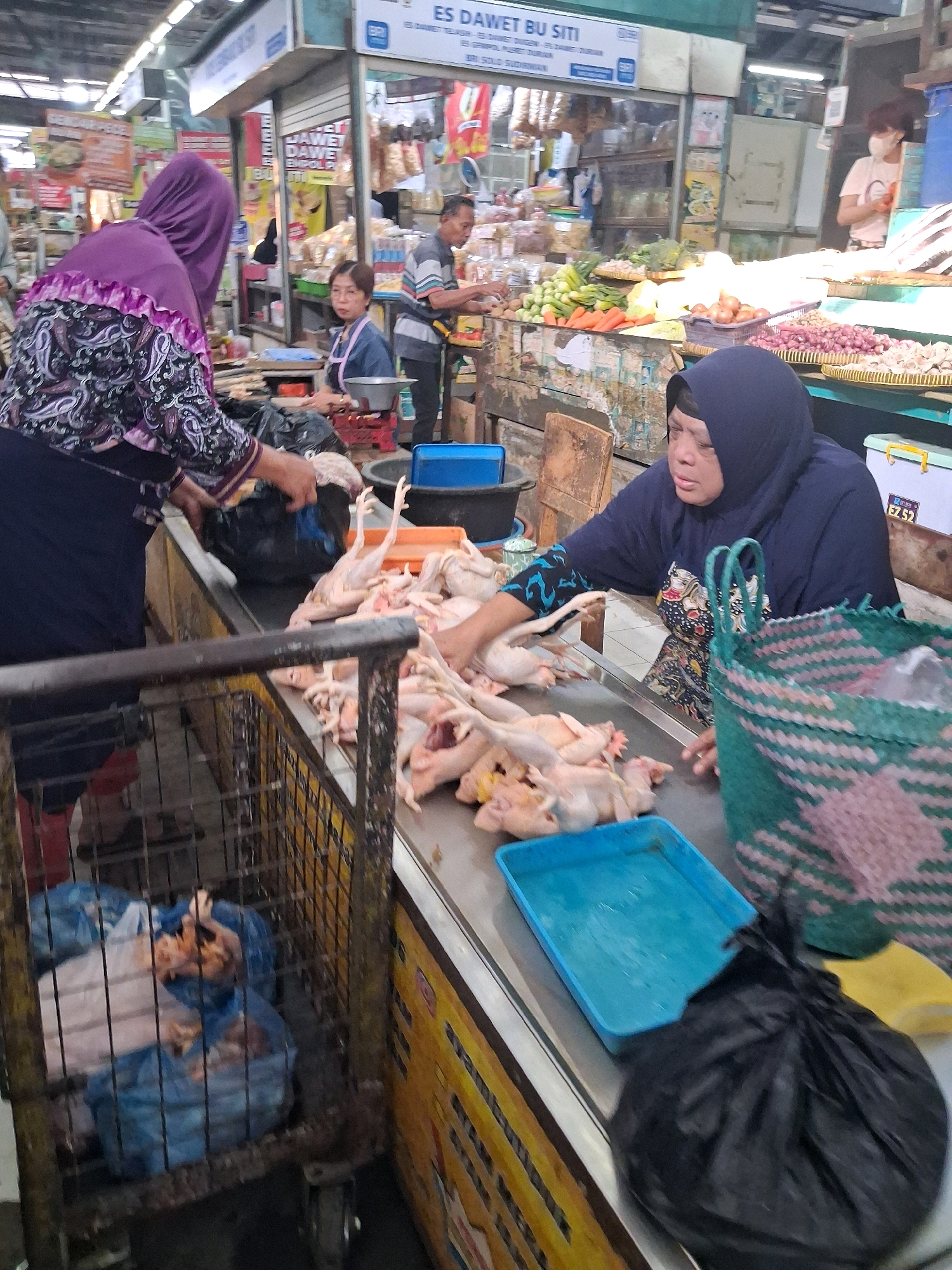 The image depicts a busy market scene in Surakarta, Indonesia. In the foreground, a woman wearing a dark blue headscarf and a patterned dress is attending to a display of raw chickens on a counter. She appears to be handling the chickens, possibly preparing them for sale. Beside her, a green woven basket is placed on the counter, and a blue plastic tray is also visible. To the left, another person wearing a purple headscarf and a patterned top is seen partially in the frame, interacting with a black plastic bag that contains more of the chickens.
Behind the main subjects, the market environment is evident with stalls and other vendors. A cage on wheels, containing several blue plastic bags filled with items, is positioned to the left of the frame. In the background, other individuals can be seen engaged in market activities, including a man in a striped shirt and a woman wearing glasses and an apron. Various products, such as vegetables, onions, and other food items, are displayed on stalls in the background. Signs with Indonesian text are visible above the stalls, indicating the names of businesses or products. The lighting suggests an indoor or covered market setting.