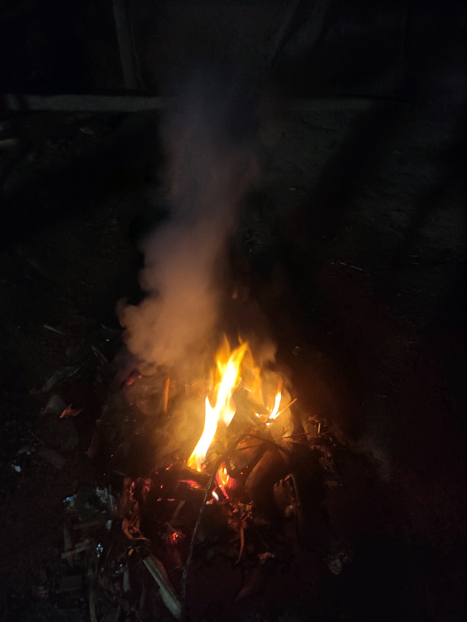 A nighttime outdoor scene in Lhokseumawe, Indonesia, shows a small bonfire with flames reaching upwards and emitting smoke. The fire is contained within a pile of debris on the ground, which appears to be composed of twigs, leaves, and other organic matter. The surrounding area is dark, with the light from the fire casting a warm glow and creating shadows. The ground surface is uneven and appears to be dirt or soil, with some scattered foliage. No individuals are visible in the image. The composition suggests a private or less populated area, possibly a backyard or a secluded spot.