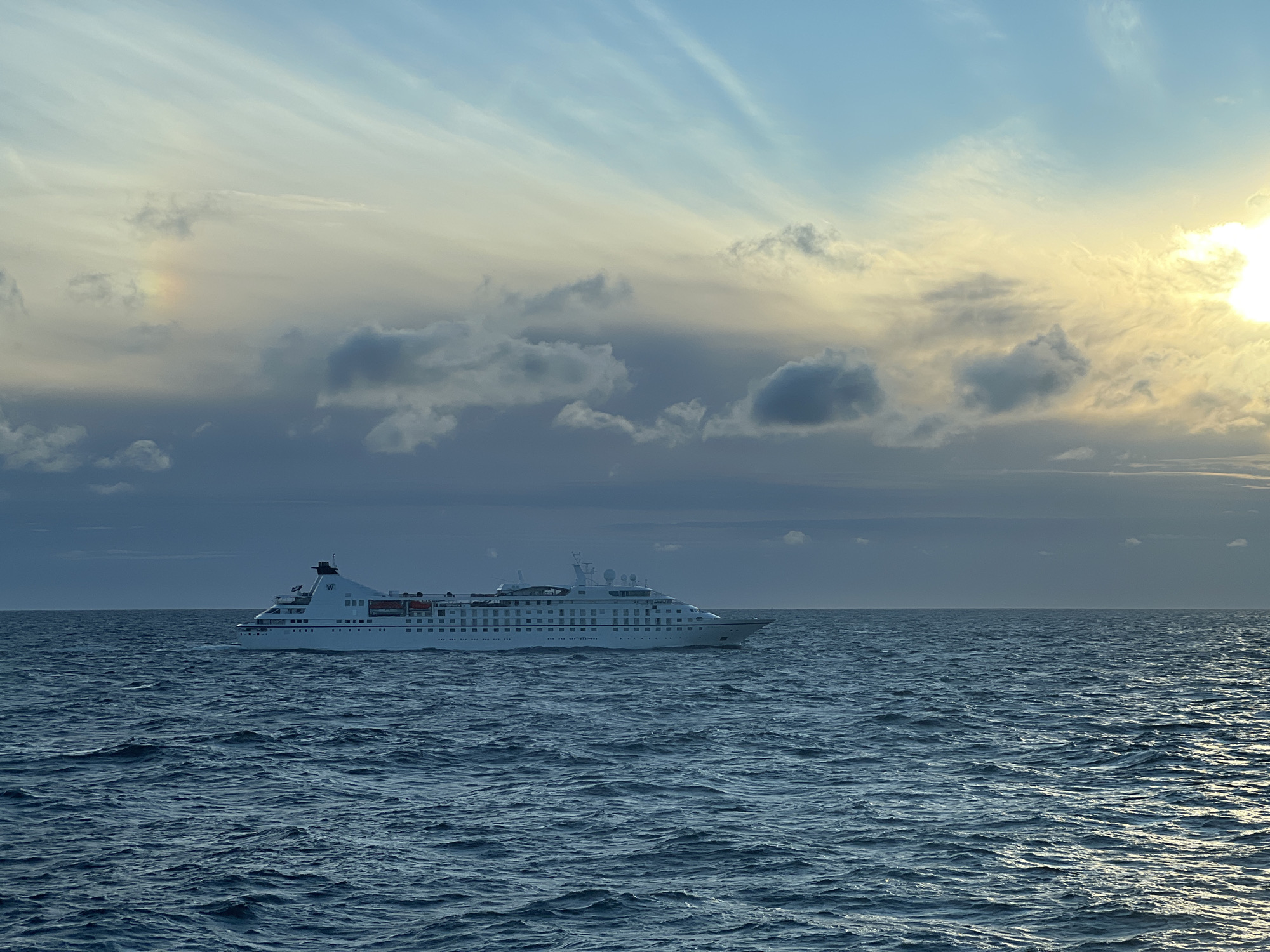 An exterior, eye-level, medium shot captures a white cruise ship on the water under a sky with clouds and the sun. The cruise ship, a single, large object, is positioned on the right side of the frame and is sailing to the left. The ship is a pale white color with multiple decks and rows of windows. The ocean fills the bottom half of the frame with choppy, dark blue water. The sky above is a gradient of colors, with lighter blues and yellows near the horizon, transitioning to darker blues at the top of the frame. There are wispy white clouds and some darker, denser clouds present in the sky. A faint rainbow-like halo is visible in the sky on the left side of the frame. No people are visible in the image. The location is unknown.