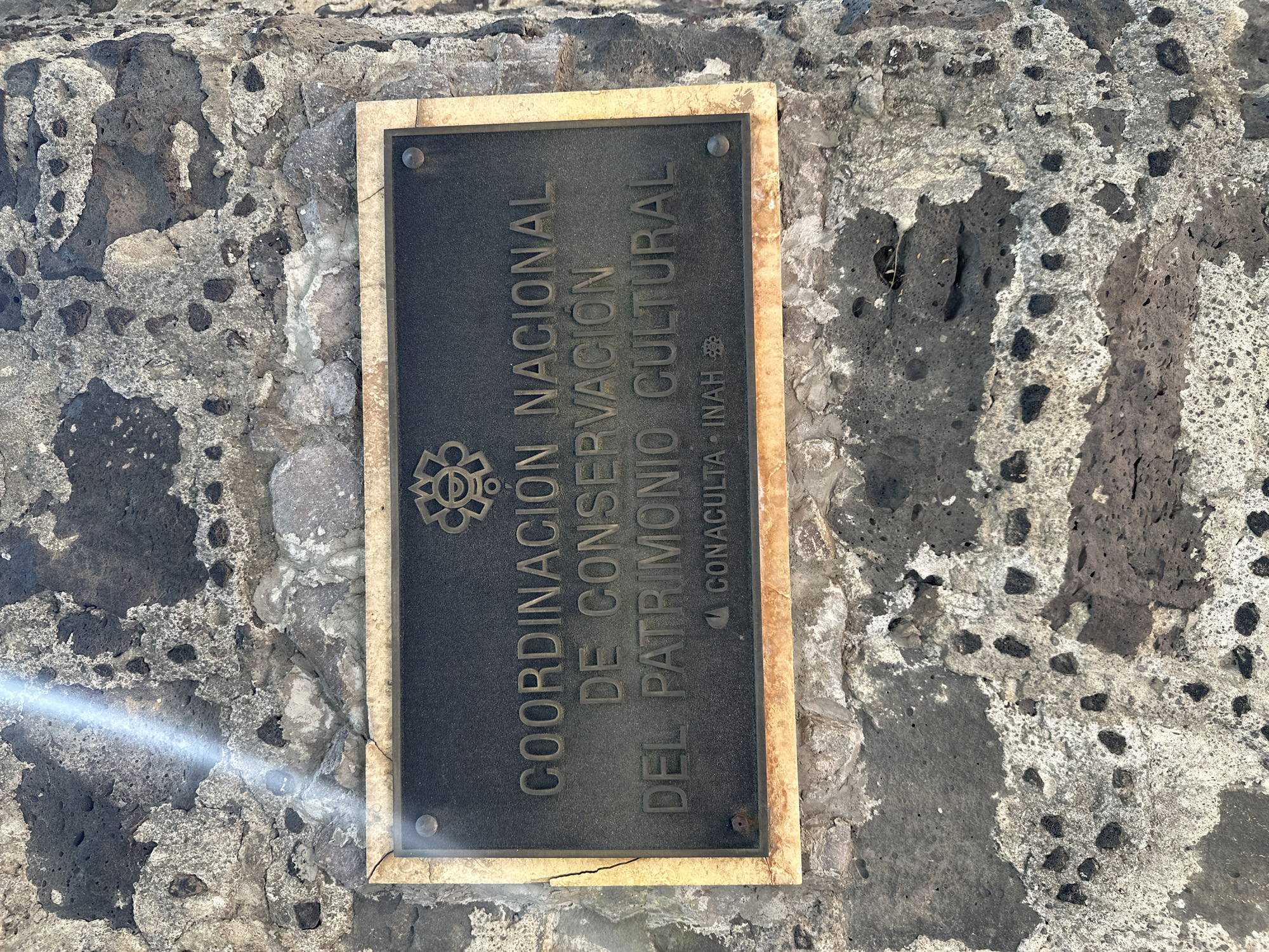The image features a dark, rectangular metal plaque mounted vertically on a textured stone wall, likely made of volcanic rock. The plaque has a light-colored, possibly stone or marble, frame, with a small crack visible on its lower left side. Four rivets secure the plaque to the wall, one visible in each corner.
Bright, direct sunlight illuminates the scene, casting a shadow from the plaque onto the wall and causing a horizontal lens flare artifact across the lower-left portion of the image, partially obscuring the wall and the plaque's frame.
The wall itself is composed of irregularly shaped, porous dark grey and lighter grey/white stones, with visible mortar filling the gaps.
The text on the plaque is in raised, capitalized Spanish letters and reads, from top to bottom:
"COORDINACIÓN NACIONAL"
"DE CONSERVACIÓN"
"DEL PATRIMONIO CULTURAL"
"CONACULTA • INAH"
Above the first line of text, a circular, intricate emblem or logo is present. The context provided indicates the location is Mexico City, Mexico, which aligns with the Spanish language text and the appearance of the volcanic stone wall. The image captures a static informational display; no activity or people are depicted.