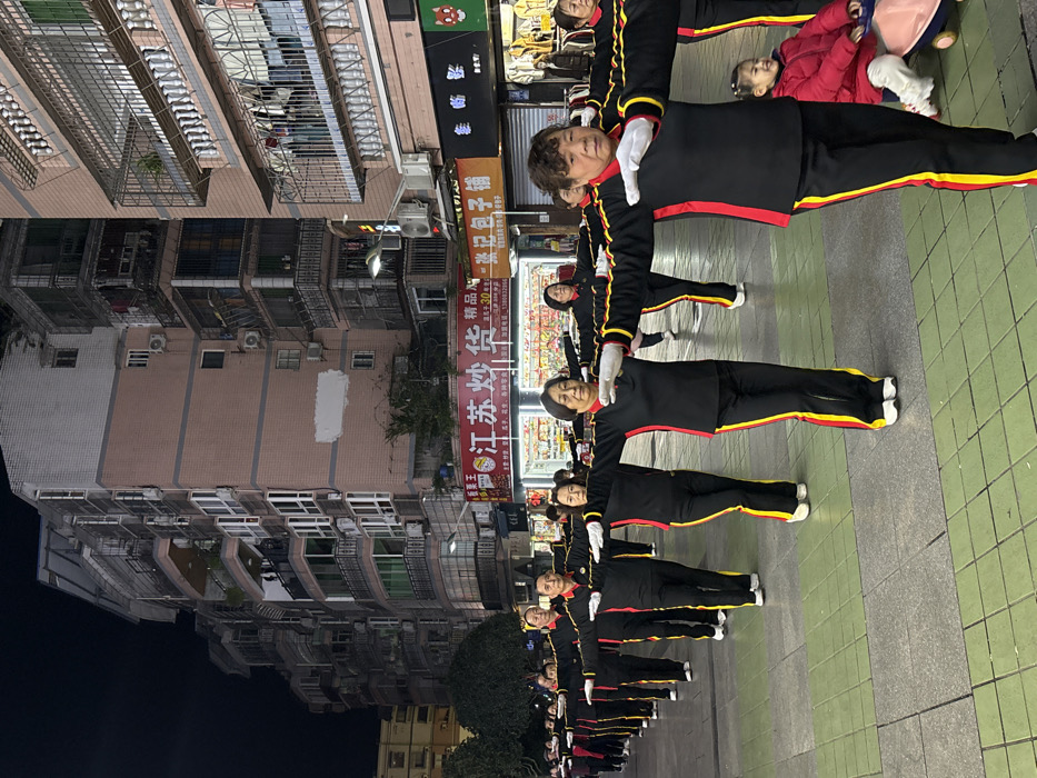 The image depicts an outdoor urban scene at night in the city of Jiangjin, China. A group of approximately 15-20 adults, primarily women, stand in a line on a tiled pavement, performing synchronized exercise with arms extended horizontally at shoulder height, wearing white gloves. Their uniform consists of black long-sleeved shirts with vertical red and yellow stripes on the sides, black pants with matching stripes, and white athletic shoes. In the immediate background, multi-story residential or mixed-use buildings with multiple windows, balconies, and air conditioning units line the street. Ground-level commercial storefronts, including one labeled "江苏炒货" (Jiangsu Fried Goods) and another "徐记包子铺" (Xu Ji Steamed Buns), are illuminated. The paved surface features a pattern of alternating grey and green rectangular tiles. A small child in a red jacket and white pants is seated on a pink and blue ride-on toy in the lower right foreground, positioned behind the exercising adults, and appears to be smiling towards the camera. The scene is illuminated by artificial lighting from streetlights and building facades.