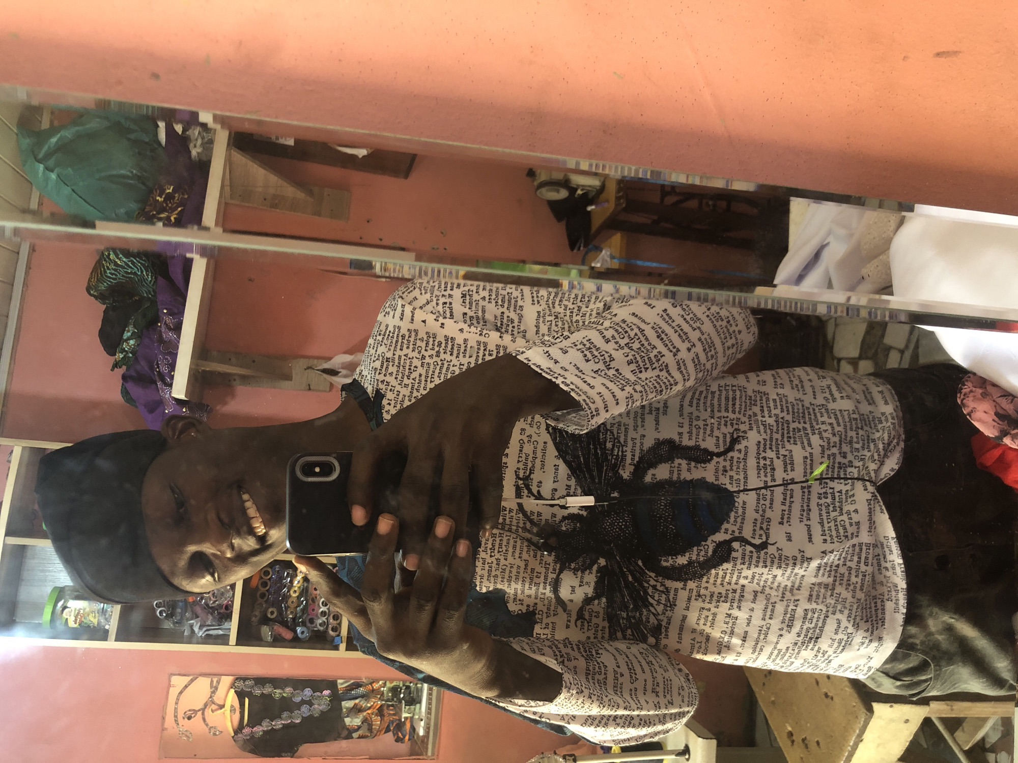 A medium shot captures a young Black man taking a selfie in a room with pink walls. He is wearing a black kufi cap, a long-sleeved shirt with a white background and black text and an image of a bee, and dark pants. He is smiling and holding a smartphone in his left hand, angled to take the picture. His right hand is also visible, holding the phone. The background reveals shelves filled with spools of thread, fabric rolls, and some folded clothing, suggesting a tailoring or fabric shop. A mirror on the right side of the frame reflects more of the room, including a sewing machine on a table, and some green and purple fabric. A framed picture with a beaded necklace is on the left wall. The lighting appears to be from overhead or a window, casting some shadows. The overall setting suggests a workshop or a retail space for tailoring supplies. The scene is likely in a city like Ado, Nigeria, given the context provided.