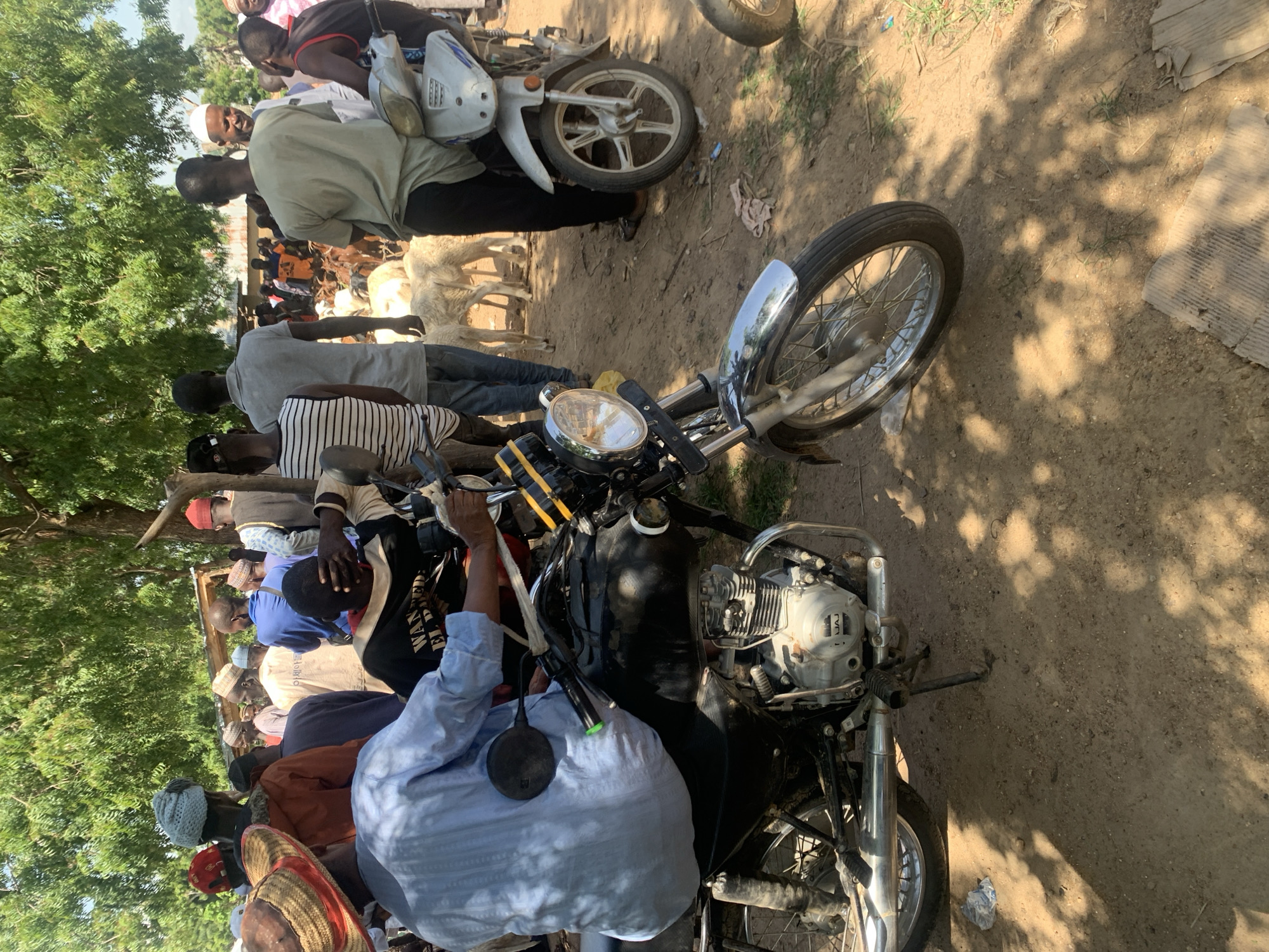 The image depicts an outdoor scene, likely a market, with a group of people and livestock. In the foreground, a motorcycle is parked, with its engine and front wheel clearly visible. A person wearing a blue shirt is seated on the motorcycle. Behind the motorcycle, several individuals are standing, some wearing traditional headwear and clothing. Sheep and goats are scattered among the people, suggesting a livestock market. The environment appears to be dusty with trees providing shade. The background shows more people and structures, hinting at a bustling gathering. The location is identified as Boi A, Nigeria.