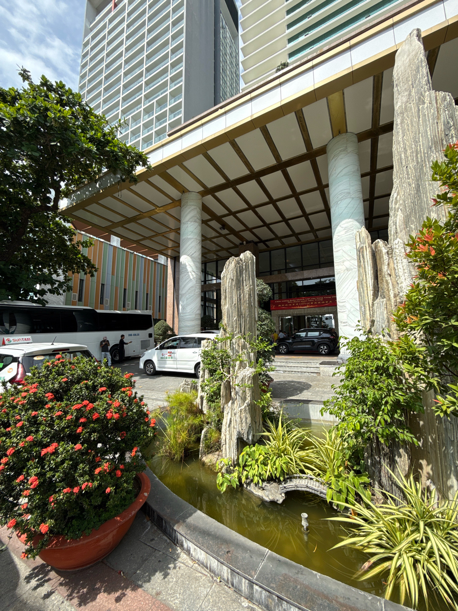 The image depicts the entrance of a hotel in Nha Trang, Vietnam, featuring a modern, multi-story building with a white facade and numerous windows. A grand entrance canopy, supported by large marble-patterned columns, is visible. The foreground is adorned with a decorative rock garden and a pond, containing water plants and two small fish. A vibrant red-flowering bush in a large terracotta pot sits to the left. Several vehicles are parked near the entrance, including a white bus with "SUNTAXI" branding, two white cars, and a black SUV. Two individuals are standing near the bus, and another person is visible through the glass doors of the hotel entrance. A red banner with Vietnamese text is displayed above the entrance. The overall scene suggests a well-maintained and inviting hotel entrance in a sunny urban environment.