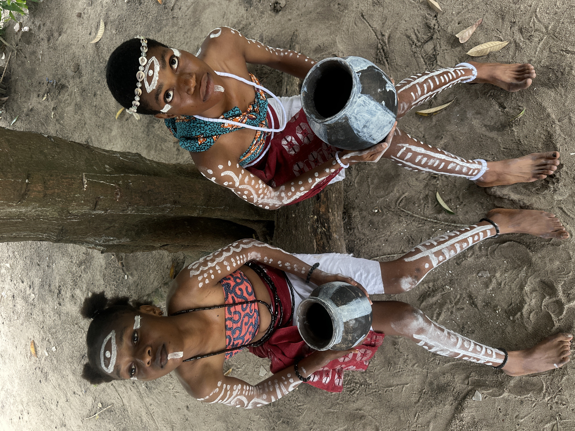 Two young individuals, adorned with white body paint and traditional attire, sit on sandy ground next to a tree in Badagry, Nigeria. The individual on the left has their hair styled in two buns and wears a beaded necklace, a patterned bandeau top, a red sash, and white shorts. They hold a dark, circular pot. The individual on the right wears a shell headband, a colorful patterned scarf, and a white necklace, and holds a similar pot. Both have white paint on their faces, arms, and legs, with decorative markings. The ground is sandy with scattered leaves, suggesting an outdoor, possibly rural or coastal, setting within Badagry. The lighting is natural, indicating it is daytime.