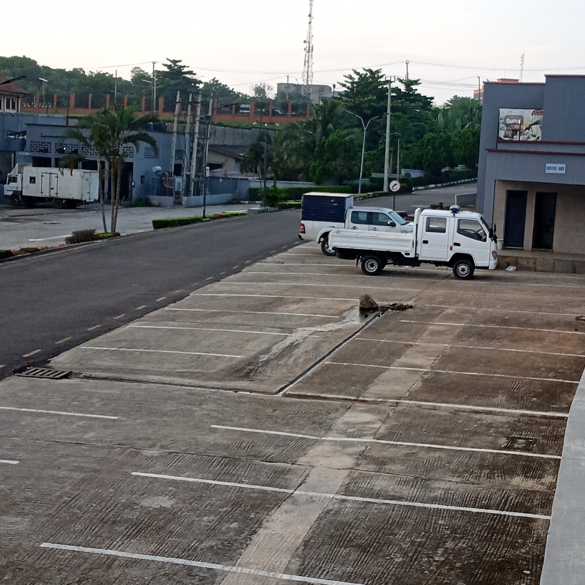 The image captures a view of a parking lot and surrounding buildings in Ibadan, Nigeria. The parking lot, made of concrete with visible expansion joints and painted white parking lines, is mostly empty. A white truck with a closed cargo bed is parked on the right side of the lot, and a white pickup truck with an open bed is parked to its left. In the background, a gray building with a sign that reads "DRIVERS SHED" is visible, along with trees and a distant cell tower. The overall scene suggests an industrial or commercial area.