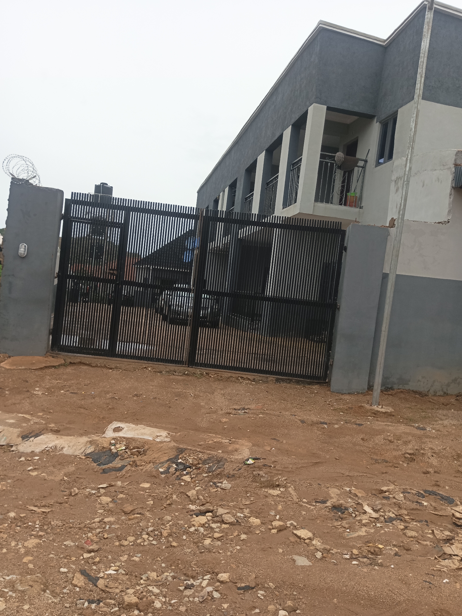 This image captures a scene in Jos, Nigeria, featuring a gated building with a modern architectural design. The building is characterized by its dark gray and light gray facade, with prominent balconies featuring black metal railings. A large, black vertical bar gate stands in the foreground, providing access to the property.
To the left of the gate, a gray pillar is visible, topped with coiled barbed wire, suggesting security measures. The ground in front of the gate and building is unpaved, consisting of reddish-brown soil mixed with stones and scattered debris, indicative of an undeveloped or construction site. The sky above is overcast, contributing to a muted and slightly dim lighting across the scene. The overall impression is one of a functional, perhaps residential or commercial, property under development or in a state of everyday use in an urban or peri-urban setting within Jos.