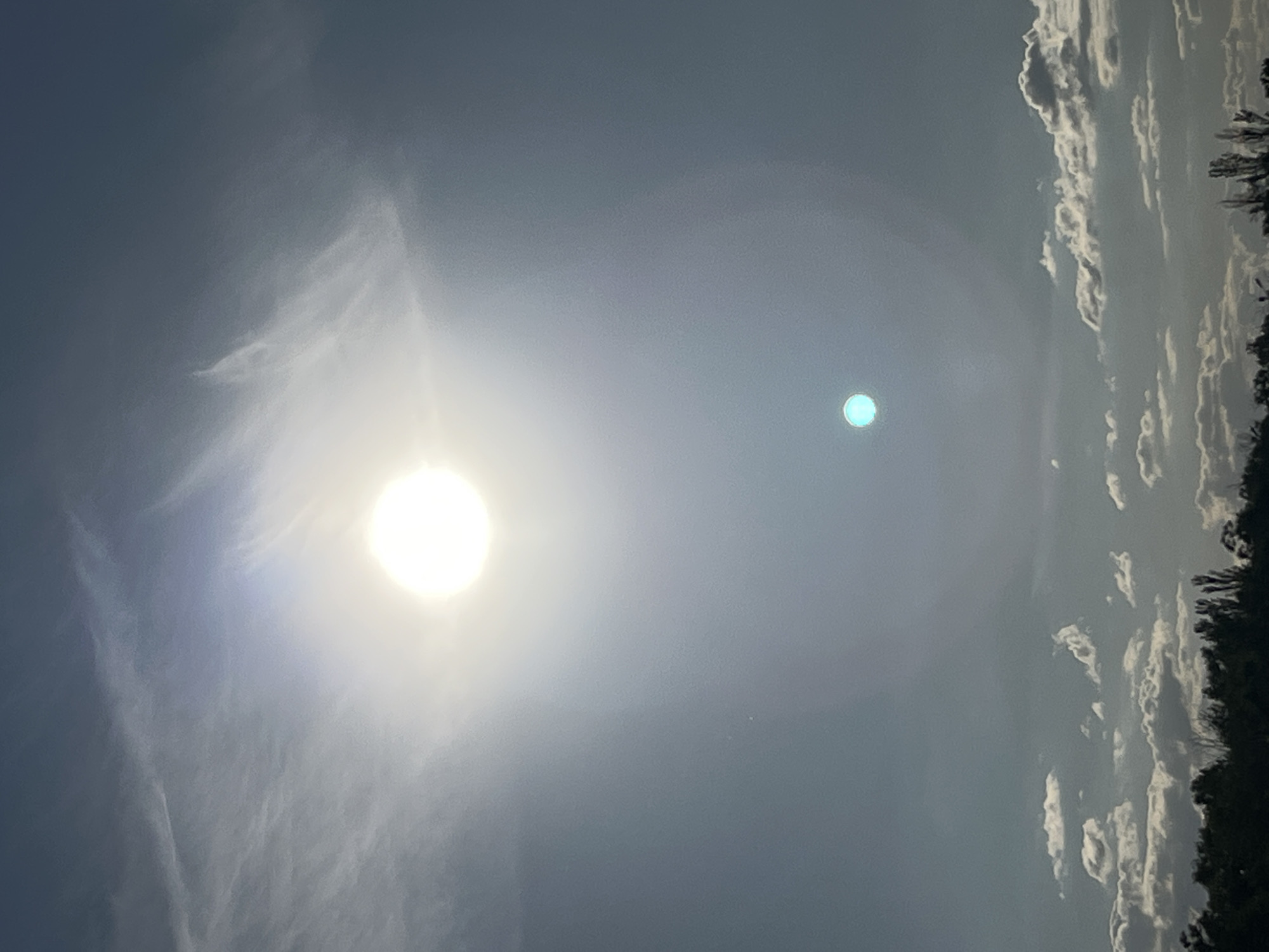 The image captures a bright sun in a partly cloudy sky, casting a lens flare that creates a secondary, smaller, aqua-colored orb below it. The scene is set against a backdrop of wispy clouds at higher altitudes and more defined cumulus clouds closer to the horizon. The bottom of the image shows a dark silhouette of trees, suggesting a natural or semi-rural environment. The overall lighting indicates it is daytime, with the sun being the primary light source. This scene is located in the vicinity of Shyroka Dacha, Ukraine.