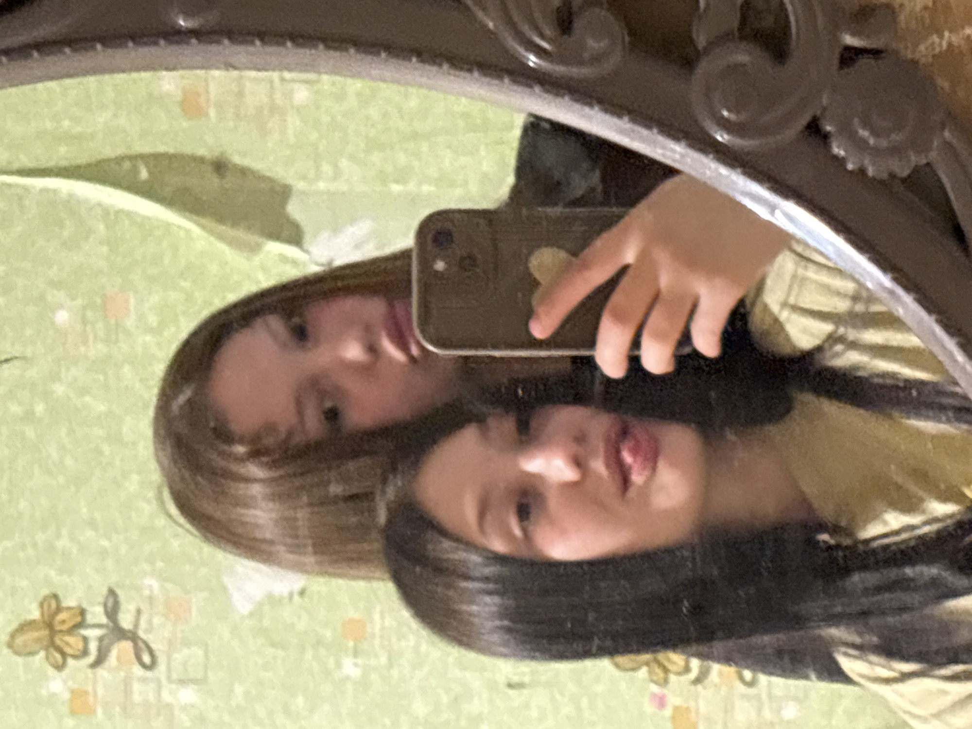 Two girls are reflected in an ornate, oval mirror, taking a selfie with a phone. The reflection also shows a light green wallpaper with floral designs and geometric patterns. The location in the city of Фоки, Russia, is not evident in the image.