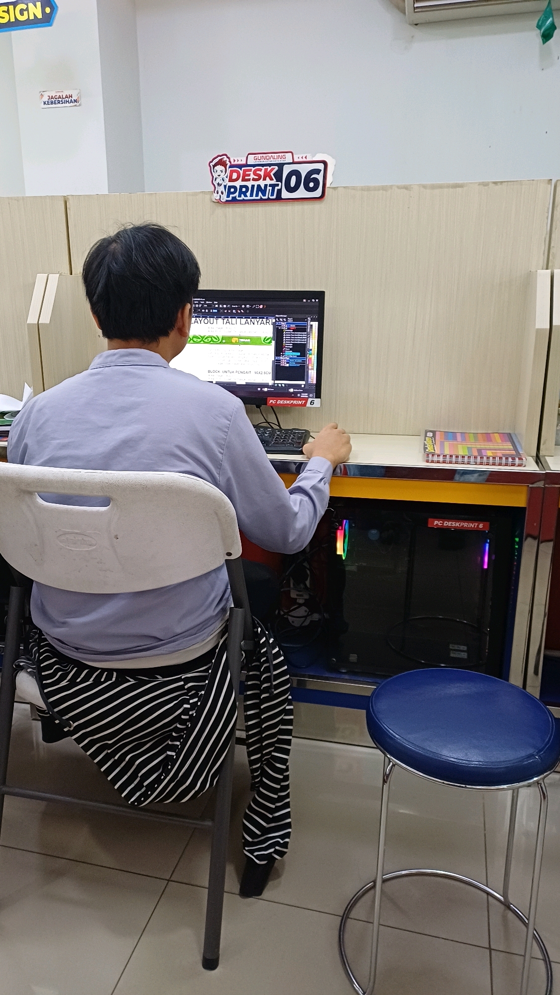 A person is seated at a computer in what appears to be a print shop or computer rental station. The person, facing away from the camera, is wearing a light blue long-sleeved shirt and black and white striped pants. The computer screen displays a design program with the words "LAYOUT TALI LANYARD" visible. A keyboard is in front of the monitor. To the right of the monitor, there is a spiral notebook with colorful covers. Below the desk, a desktop computer tower is visible with multicolored LED lights. A blue stool with a chrome base is positioned to the right of the chair. Above the desk, a sign with "GUNDALING DESK PRINT 06" is displayed. Another sign on the wall behind the person reads "JAGALAH KEBERSIHAN" (Maintain Cleanliness). The environment appears to be an indoor space with tiled flooring and partition walls. The scene suggests a commercial setting in Cibinong, Indonesia, likely a business offering printing or computer services.