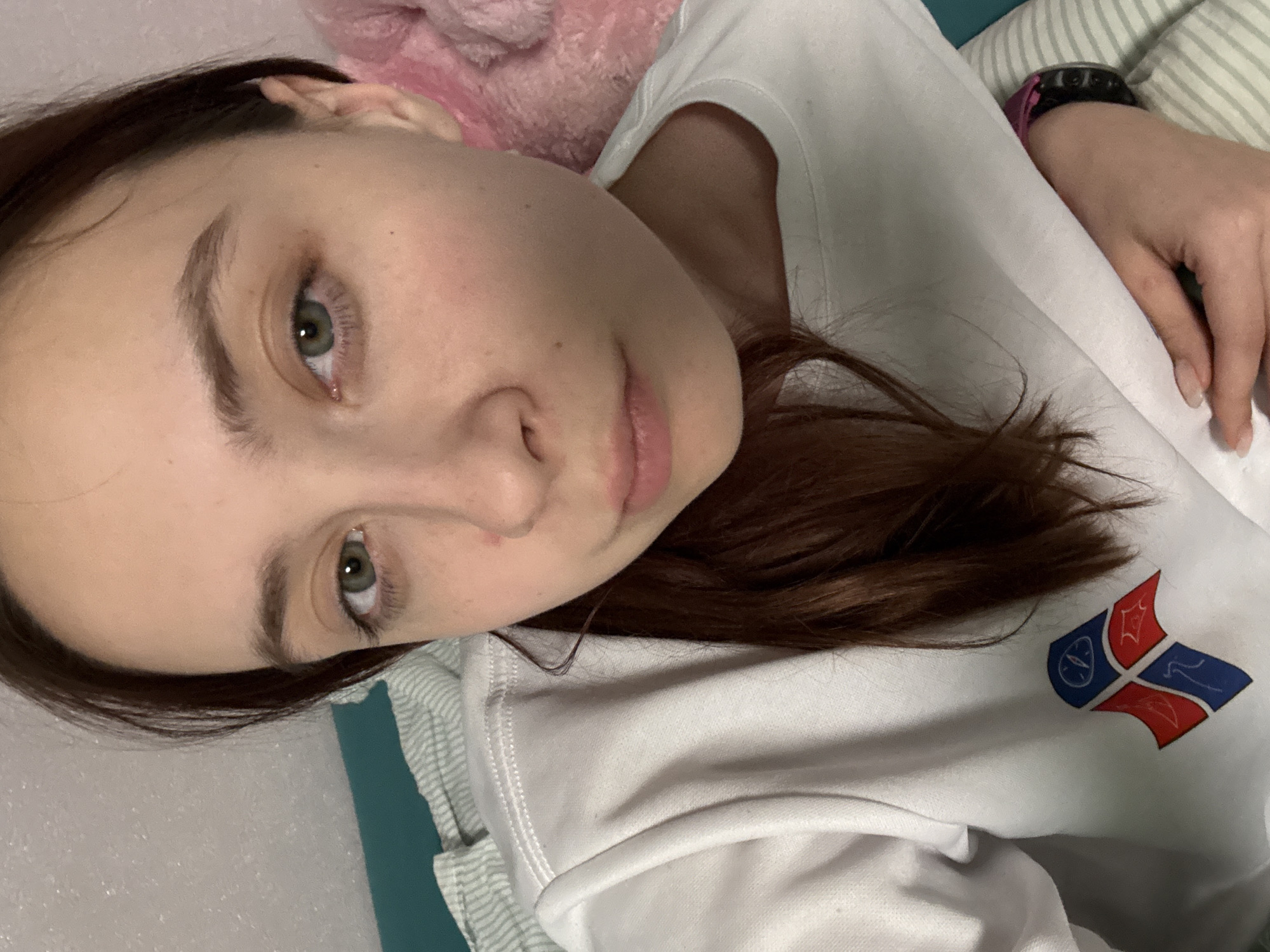 A close-up photograph features a young woman with brown hair and fair skin. She is lying down, her head tilted towards the camera. Her eyes are open, and she appears to be looking directly at the viewer. She is wearing a white shirt with a logo of what appears to be a flag on it. In the background, there is a glimpse of a striped pillow and a pink object. A black watch is visible on her wrist. The overall lighting is soft and diffused. There is no indication of the photograph being taken in the city of Прикамский, Russia, but it is likely taken inside a home.