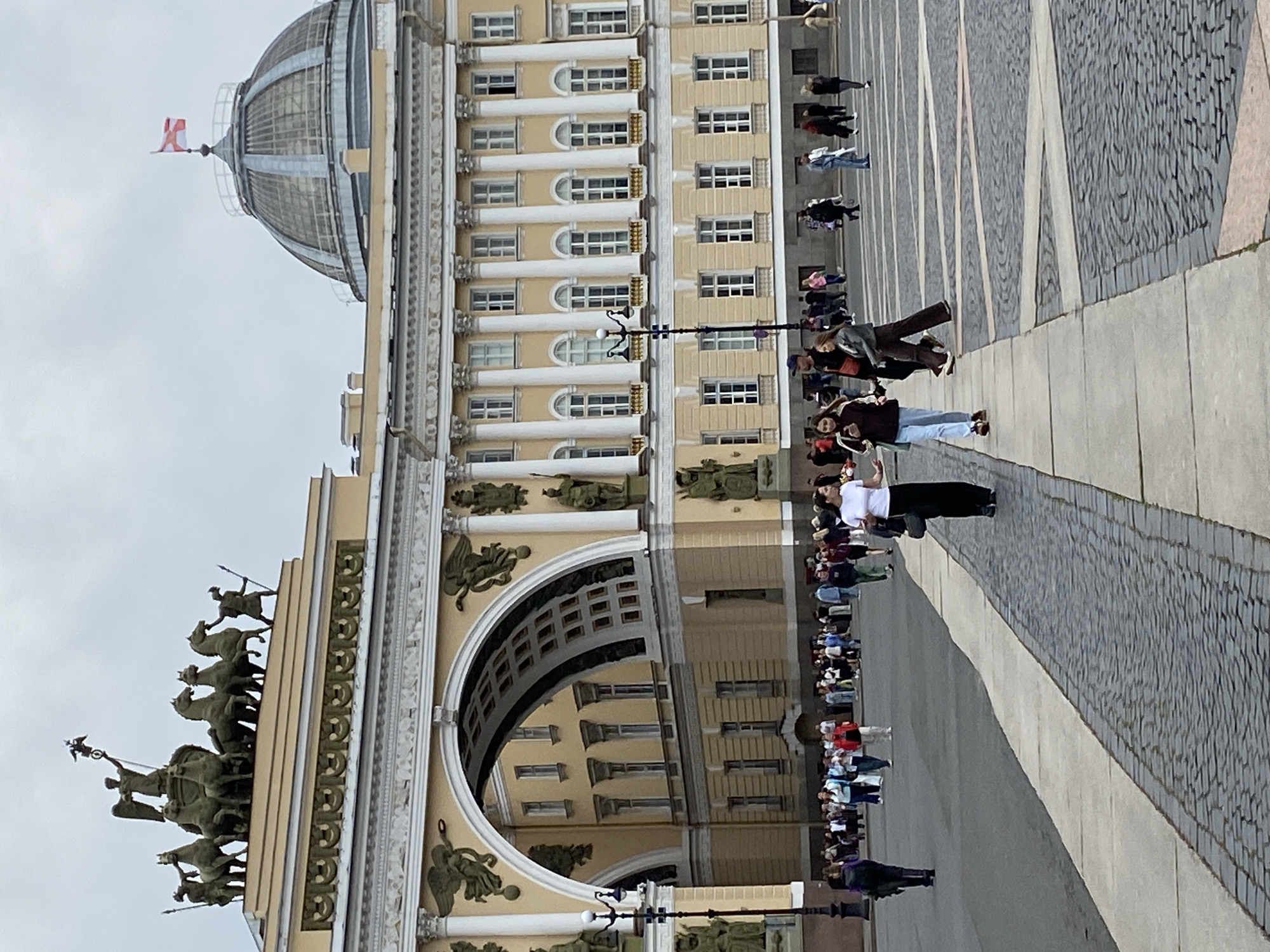 The image shows a view of Palace Square in Saint Petersburg, Russia, on an overcast day. The General Staff Building's Triumphal Arch, adorned with a chariot sculpture, dominates the left side of the frame. To the right, the dome of the Winter Palace is visible, topped with a Russian flag. In the foreground, several people are walking across the paved square. Some are dressed in casual clothing, and one woman is wearing a long black skirt. The ground is a mixture of cobblestones and paved walkways. The overall atmosphere appears to be that of a public space with moderate foot traffic.