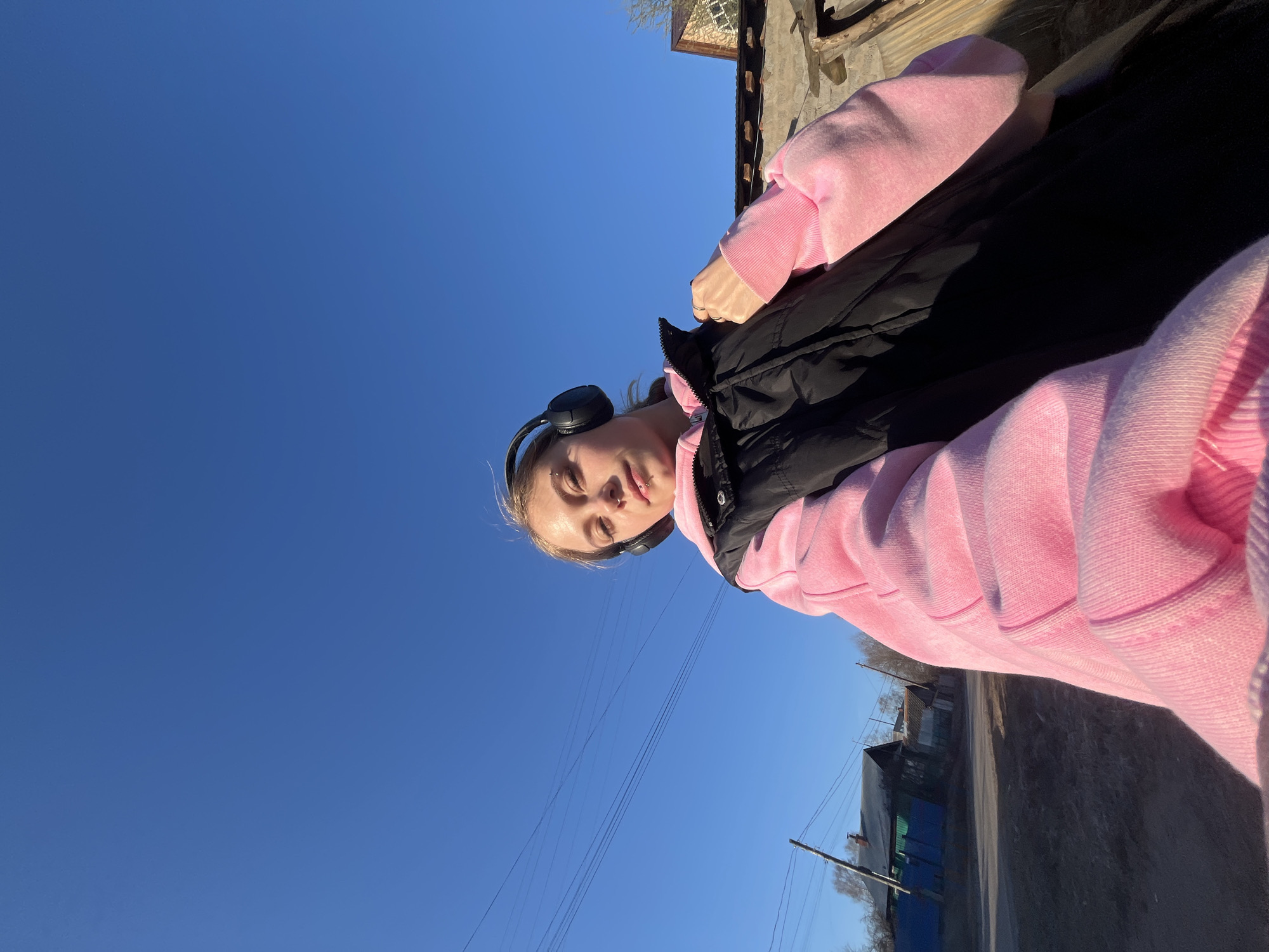 A vertically oriented outdoor selfie captures a young person in Фоки, Russia, under a brilliant, clear blue sky. The subject, a young female with light hair, is wearing black over-ear headphones. She has multiple facial piercings, including on her eyebrow, nose, and lip. She is dressed in a bright pink hooded sweatshirt, layered with a black puffer vest. A ring is visible on her right hand. Her expression is neutral, with a slight, subtle smile.
The image is taken from a low angle, looking upwards, with the vast expanse of the clear blue sky occupying a significant portion of the frame. The strong, direct sunlight casts distinct shadows, indicating a bright day, likely morning or afternoon. In the background, to the right and lower part of the image, there are sparse, leafless trees, suggesting late winter or early spring, along with utility poles and overhead wires. Some rustic buildings or structures are also visible in the distance, suggesting a rural or semi-rural setting with an unpaved road or path. No discernible text is visible.