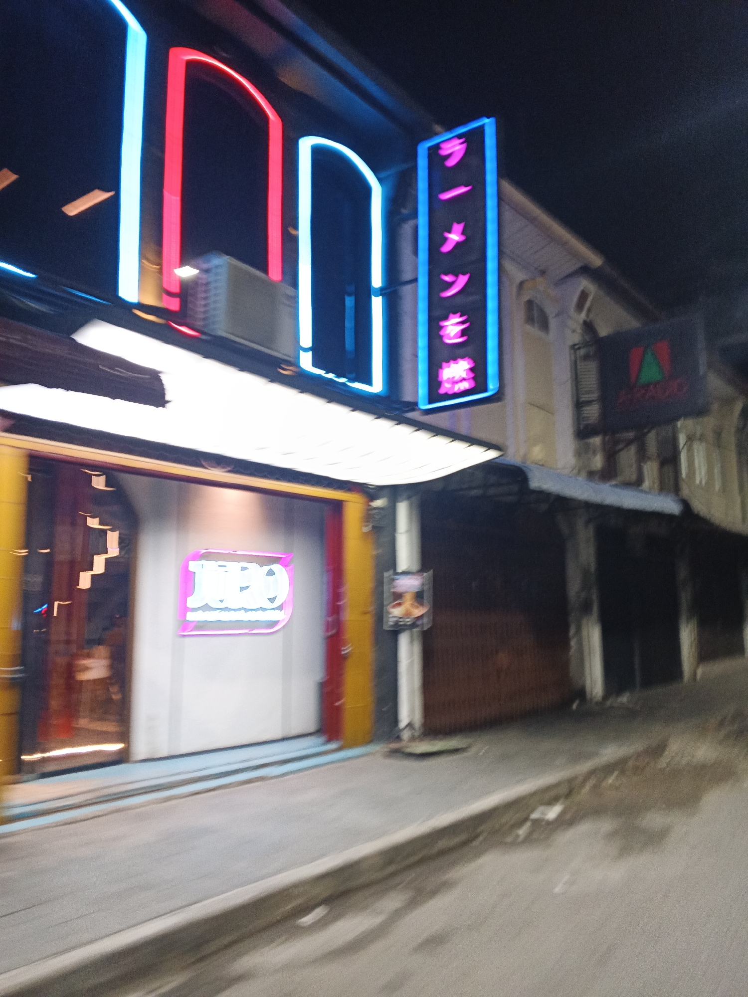 A night time, blurry, street level, full shot captures a row of businesses on the side of a road in the city of Medan, Indonesia. The businesses are adorned with bright, neon signs that illuminate the dark street. On the left, a business with yellow and red accents has a large, bright white sign that appears to say "JEO" in pink neon. To the right of this, a tall, vertical neon sign in pink letters against a blue background reads "ラーメ」を燃" in Japanese characters. Further to the right, a dark sign with a green triangle and red lettering appears to read "ARAEEO". The street itself is paved and slopes slightly downhill to the right. The overall impression is that of a vibrant nightlife area.